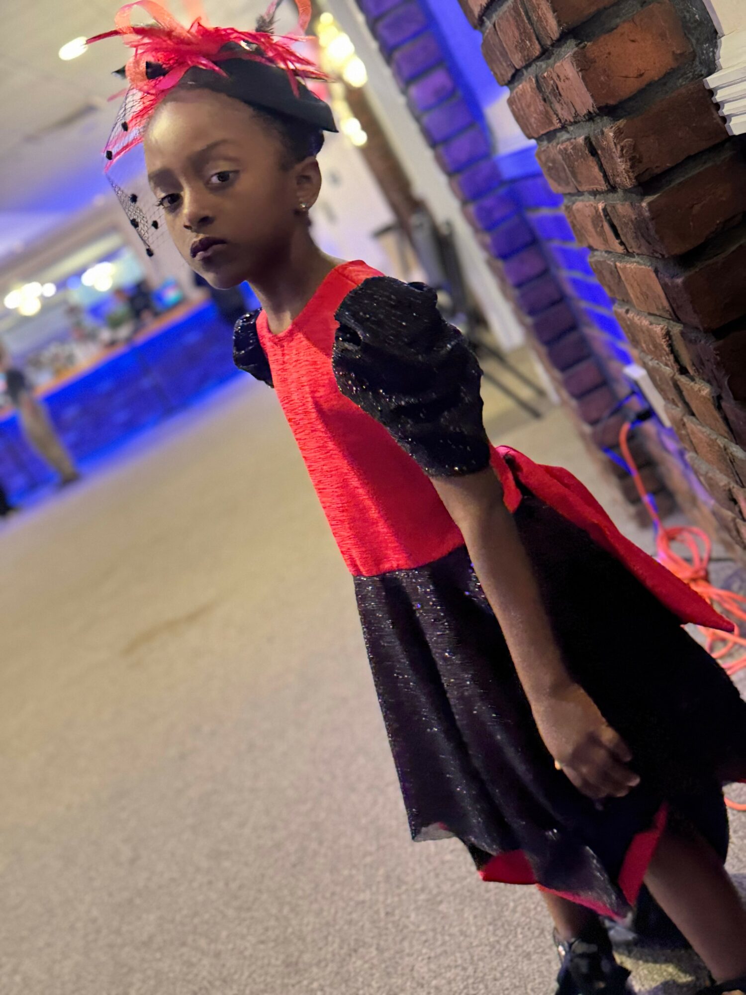 Young girl in a red and black dress with a red headband, standing near a brick wall indoors.