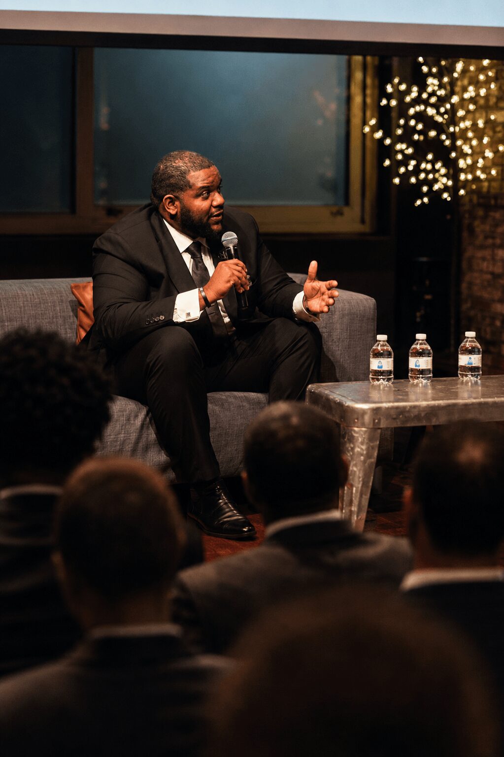 Man in suit speaking into a microphone during a panel discussion, with an audience in front, bottles on table, and decorative lights behind.