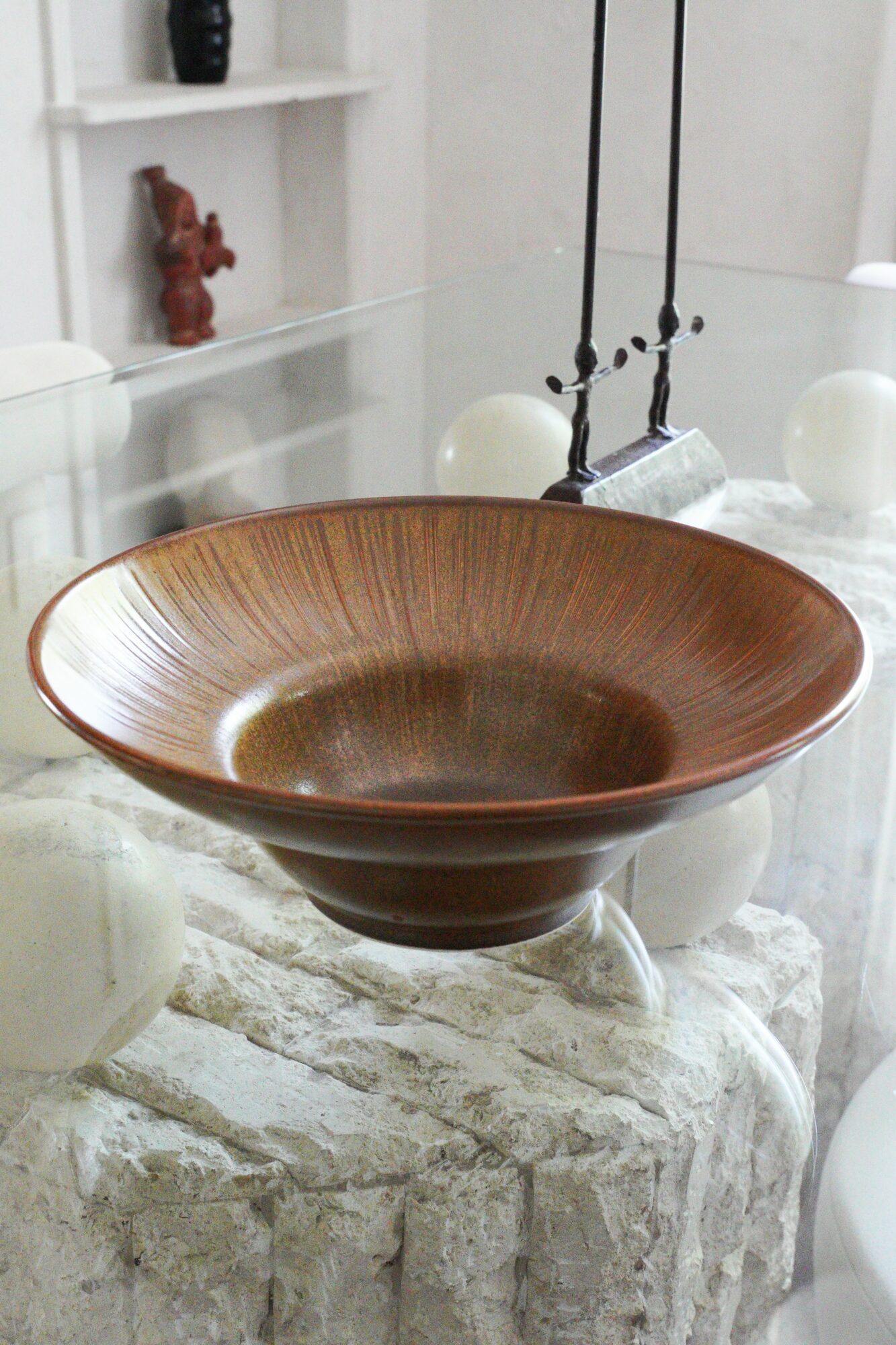 Brown ceramic bowl on a marble table with white decorative objects and a hanging light fixture in the background.