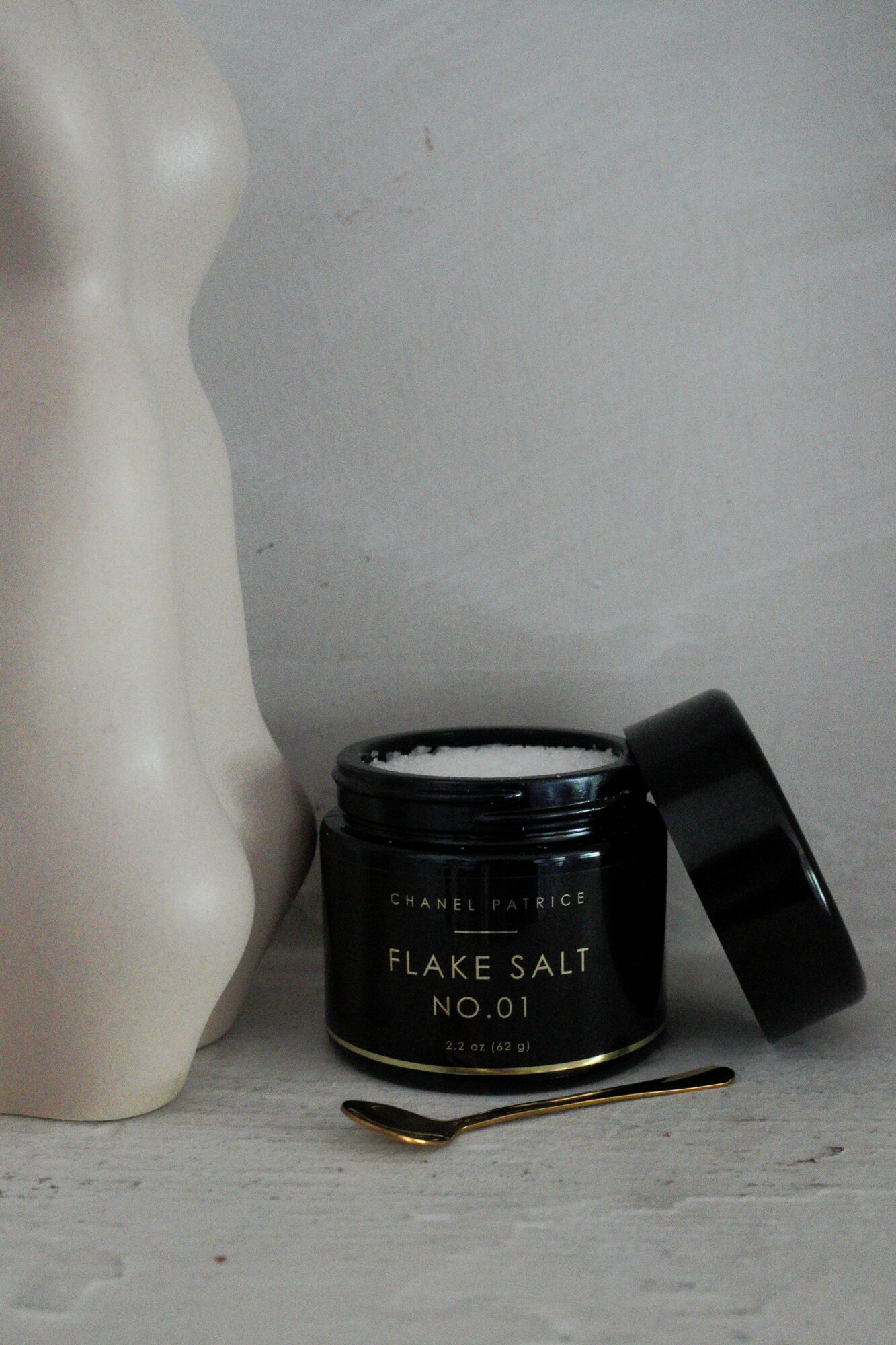 Black jar labeled 'Flake Salt No.01' with open lid, gold spoon, and mannequin torso on light gray surface.