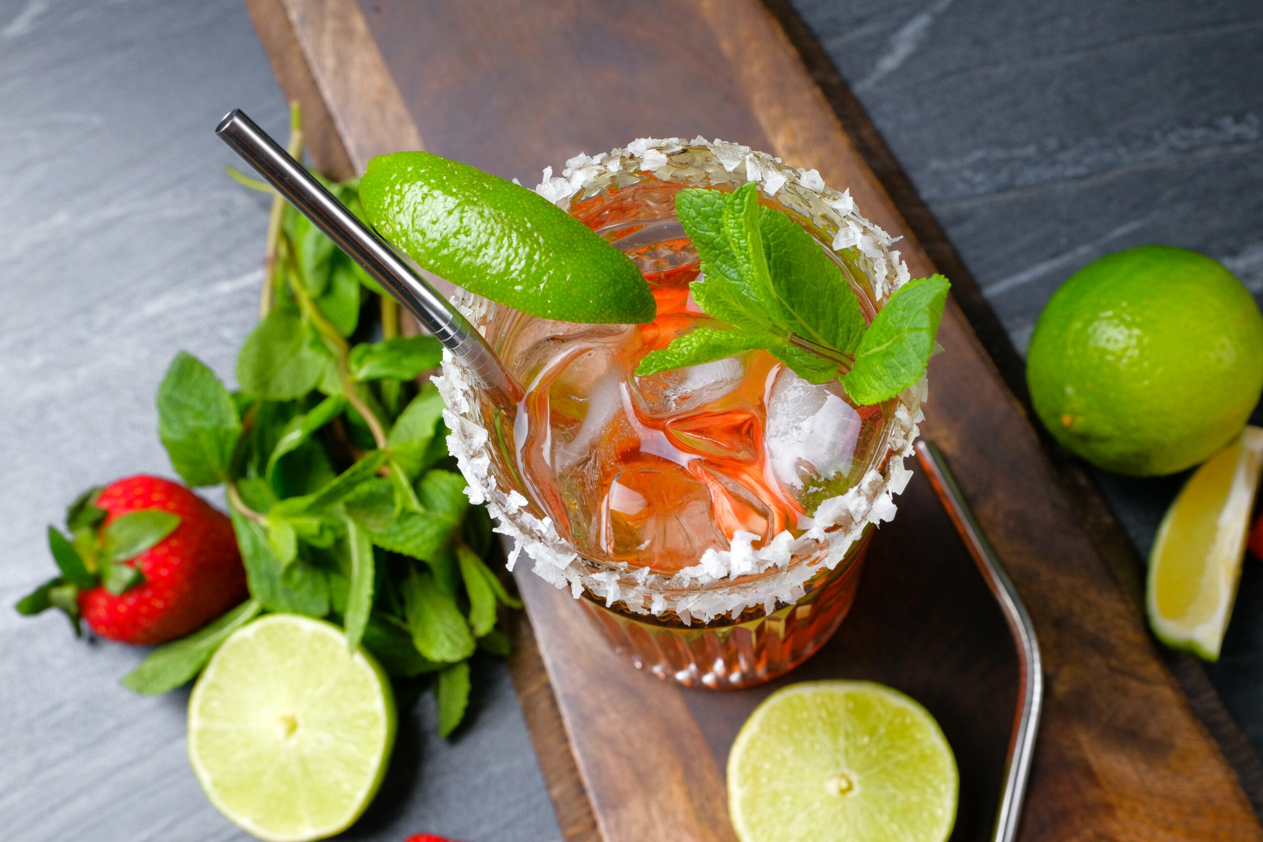 A cocktail garnished with lime, mint, and a straw, with strawberries, lime, and mint leaves nearby on a wooden surface.