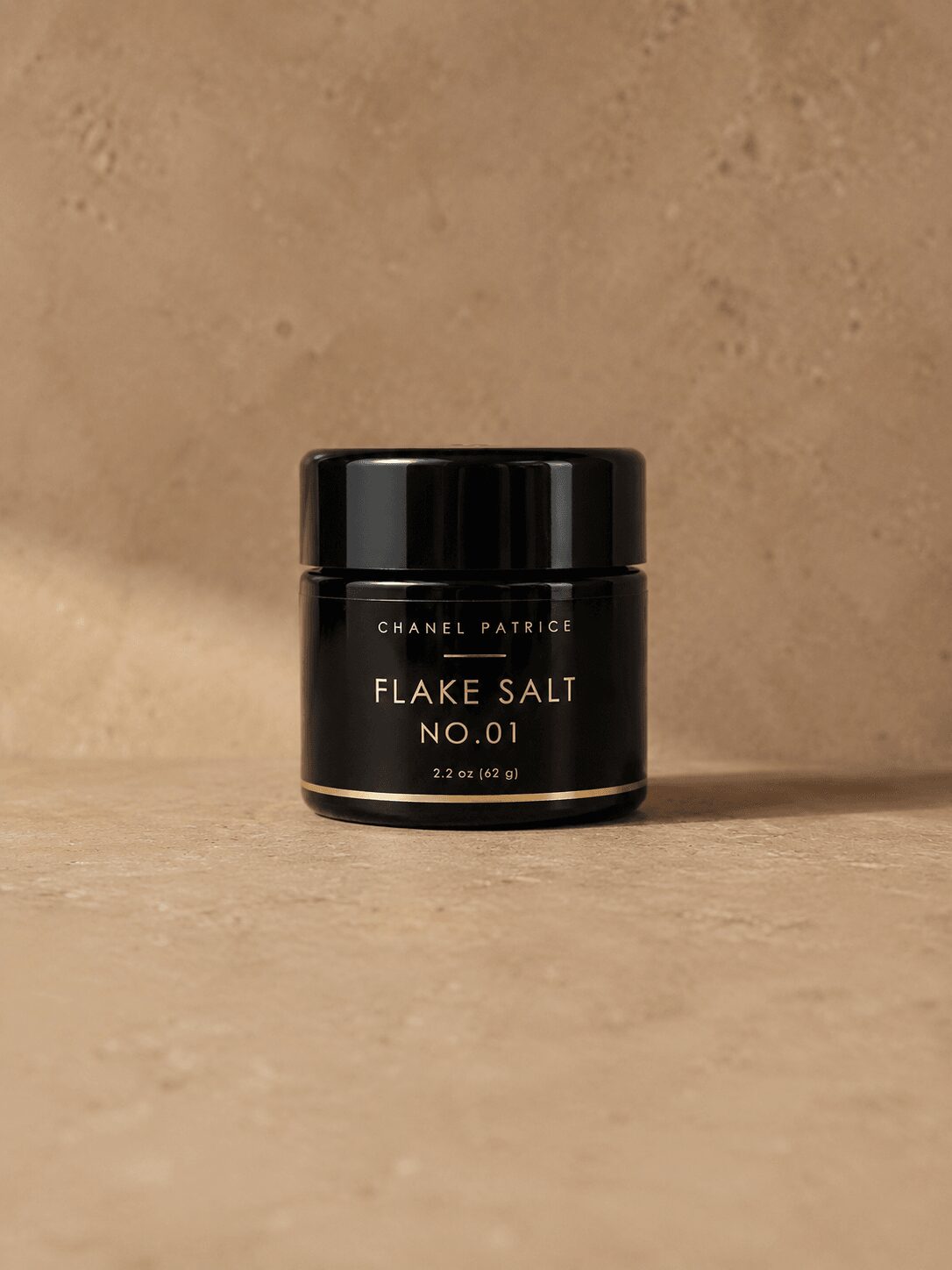 Small black jar labeled 'Chanel Patrice Flake Salt No. 01' on a beige surface with a beige background.