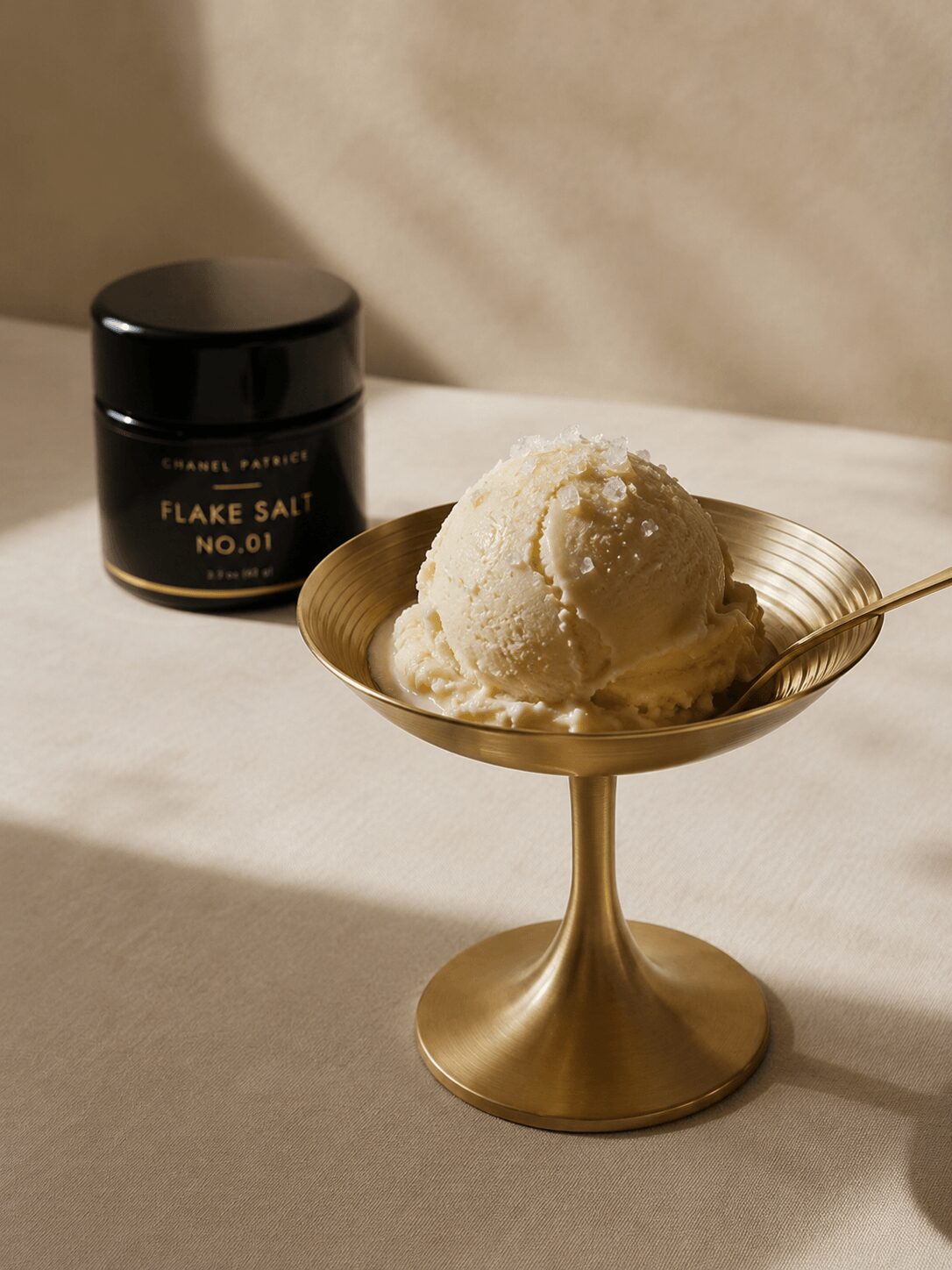 A scoop of vanilla ice cream in a gold-colored dish with a spoon, and a black jar labeled 'Flake Salt No.01' in the background.