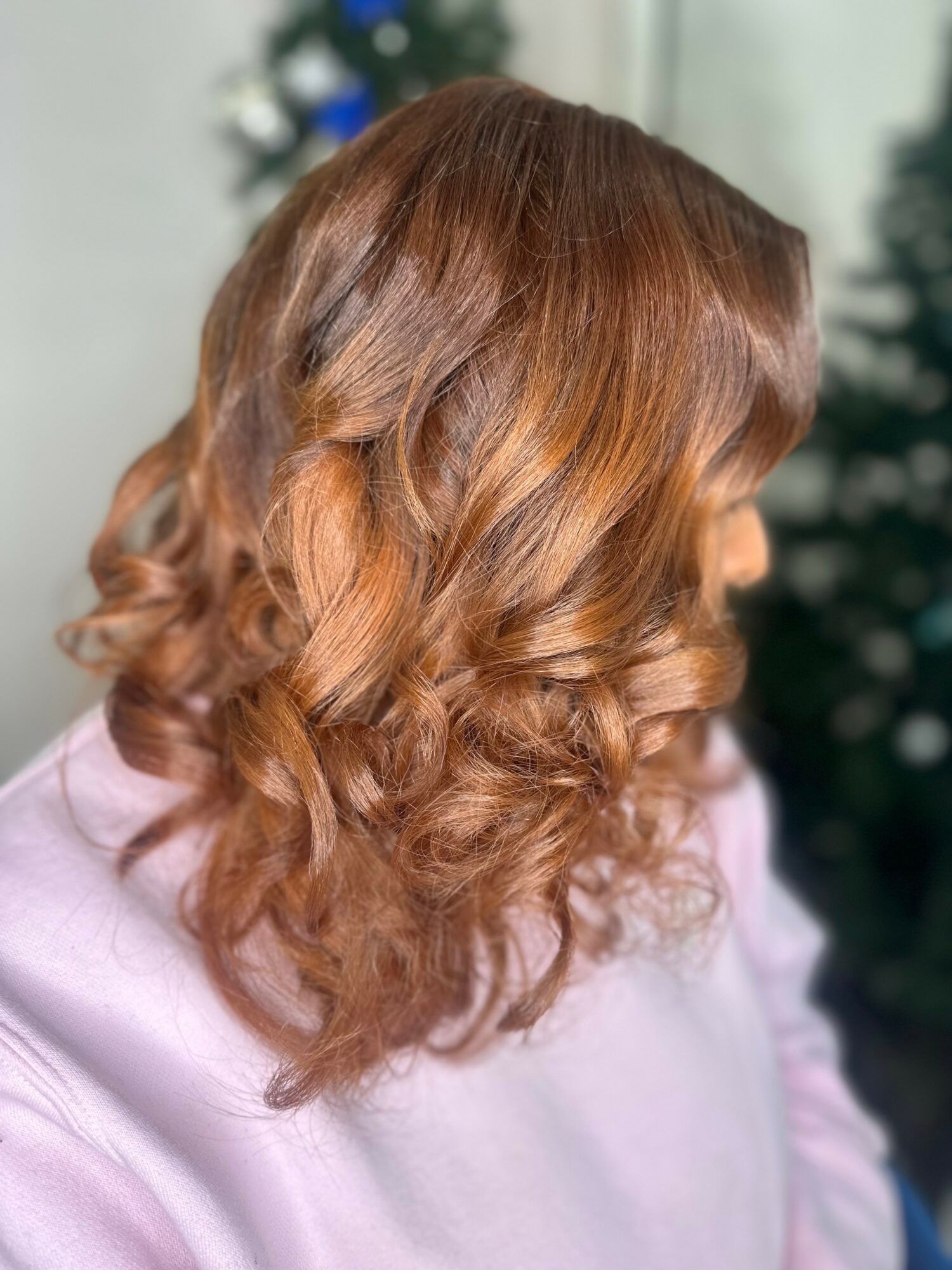 Side view of a woman with wavy, reddish-brown hair, wearing a light pink top, with a blurred background.