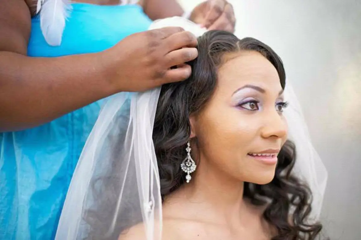 Woman with long dark hair and earrings, getting a veil placed on her head, smiling softly.