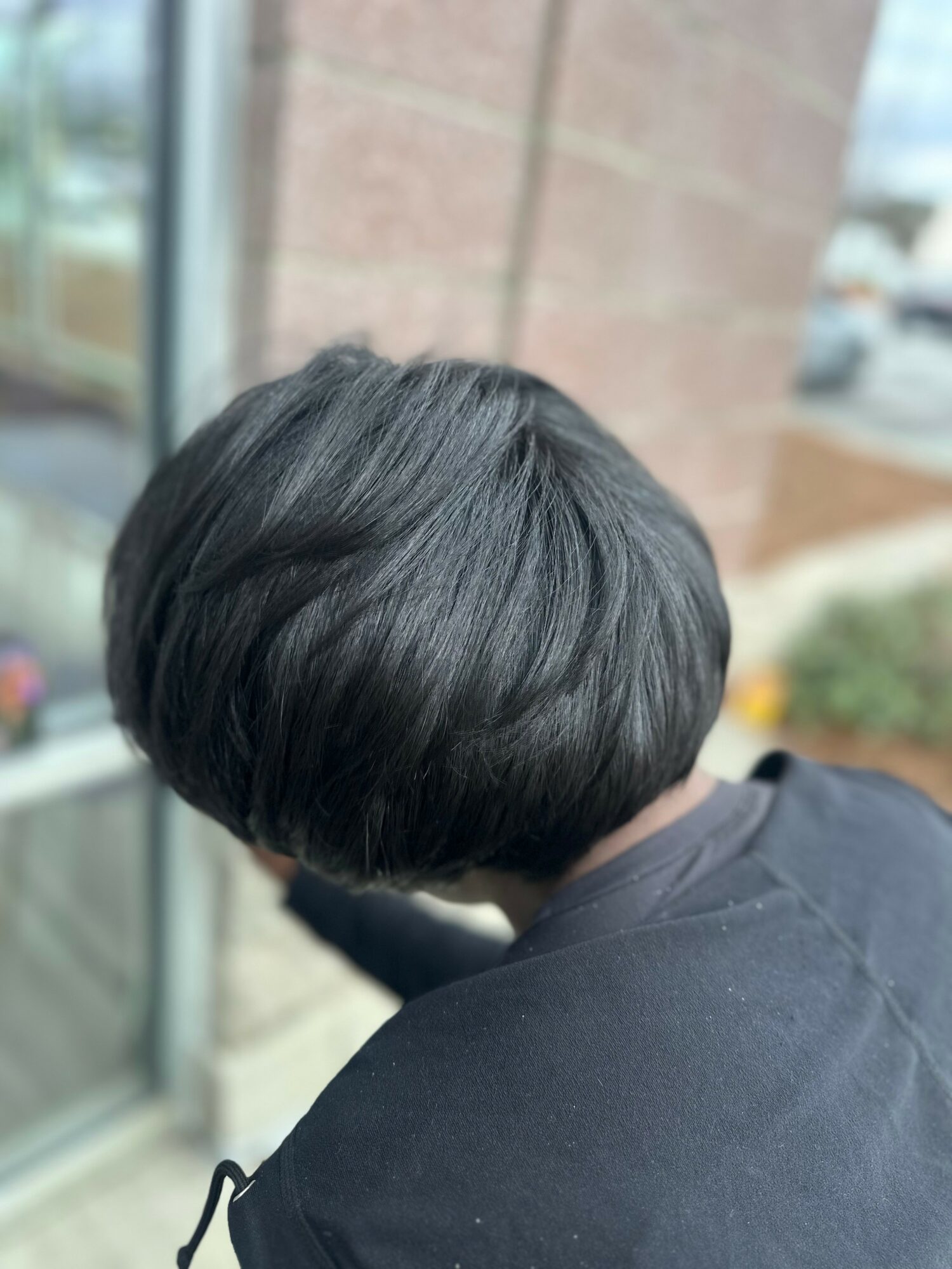 Person with short black hair, viewed from behind, outside near a window and brick wall.