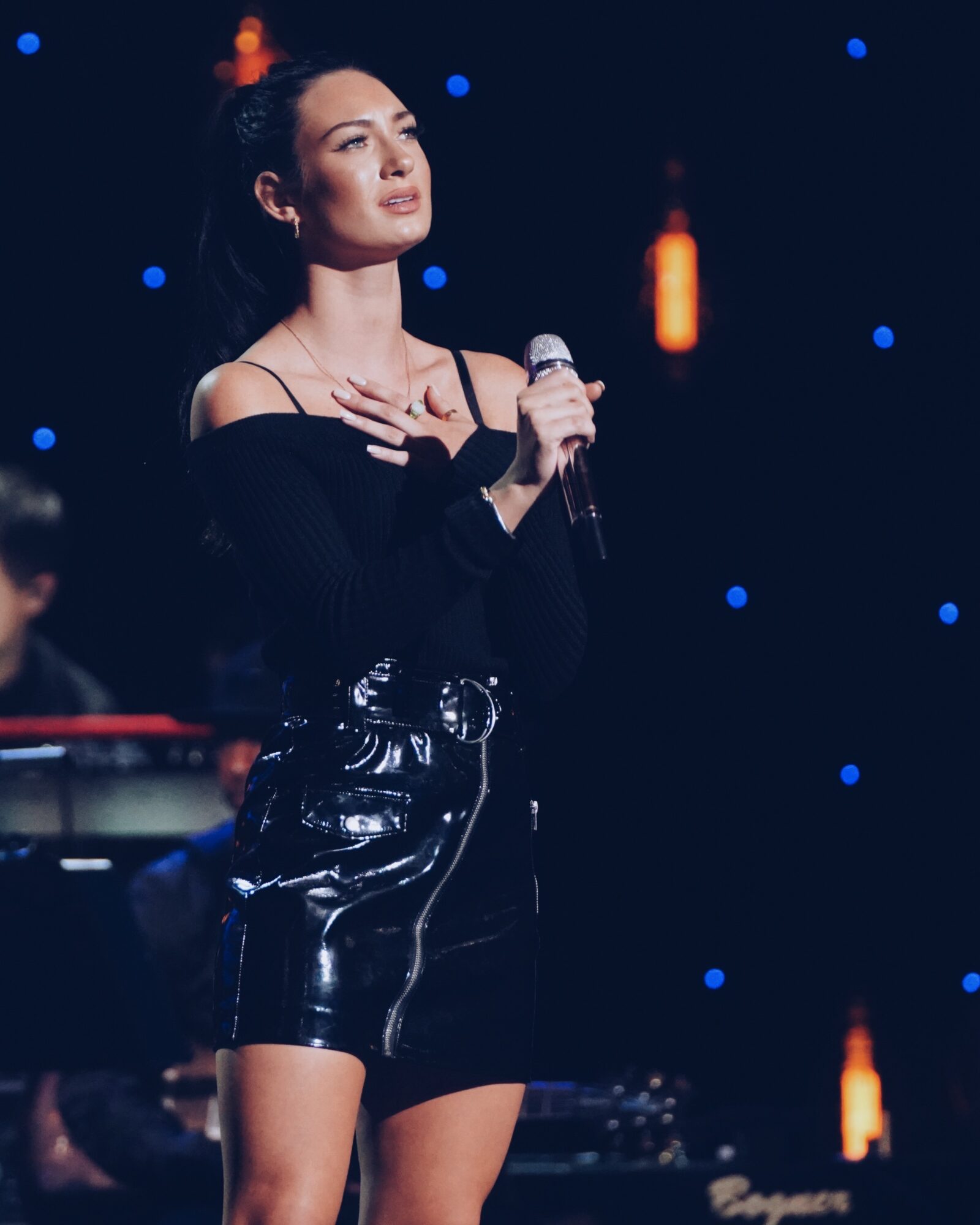 Woman holding a microphone on stage with dark background and small blue lights, wearing a black off-shoulder top and shiny shorts.