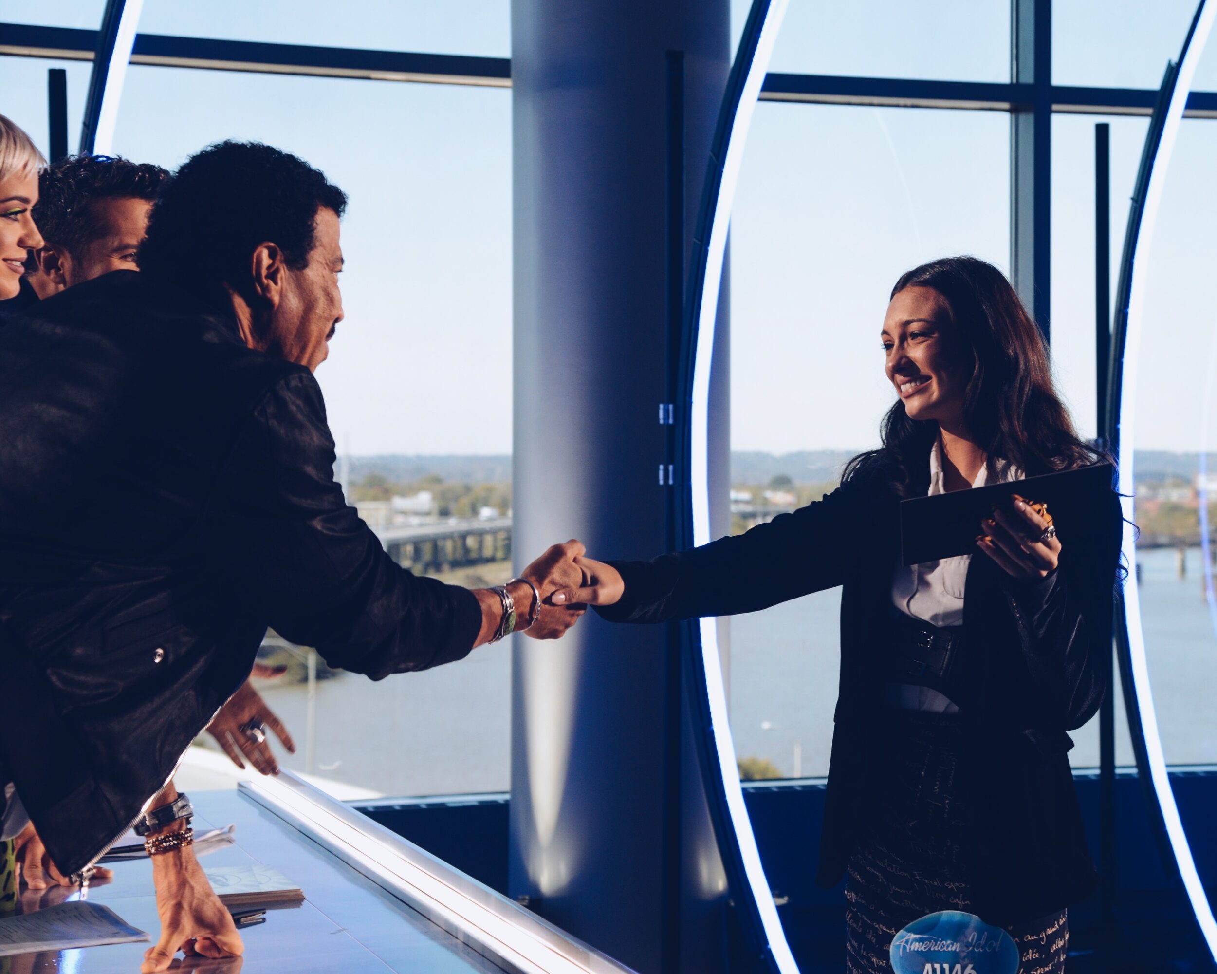 Two people shake hands in a modern office with large windows and a city view, smiling and engaging professionally.