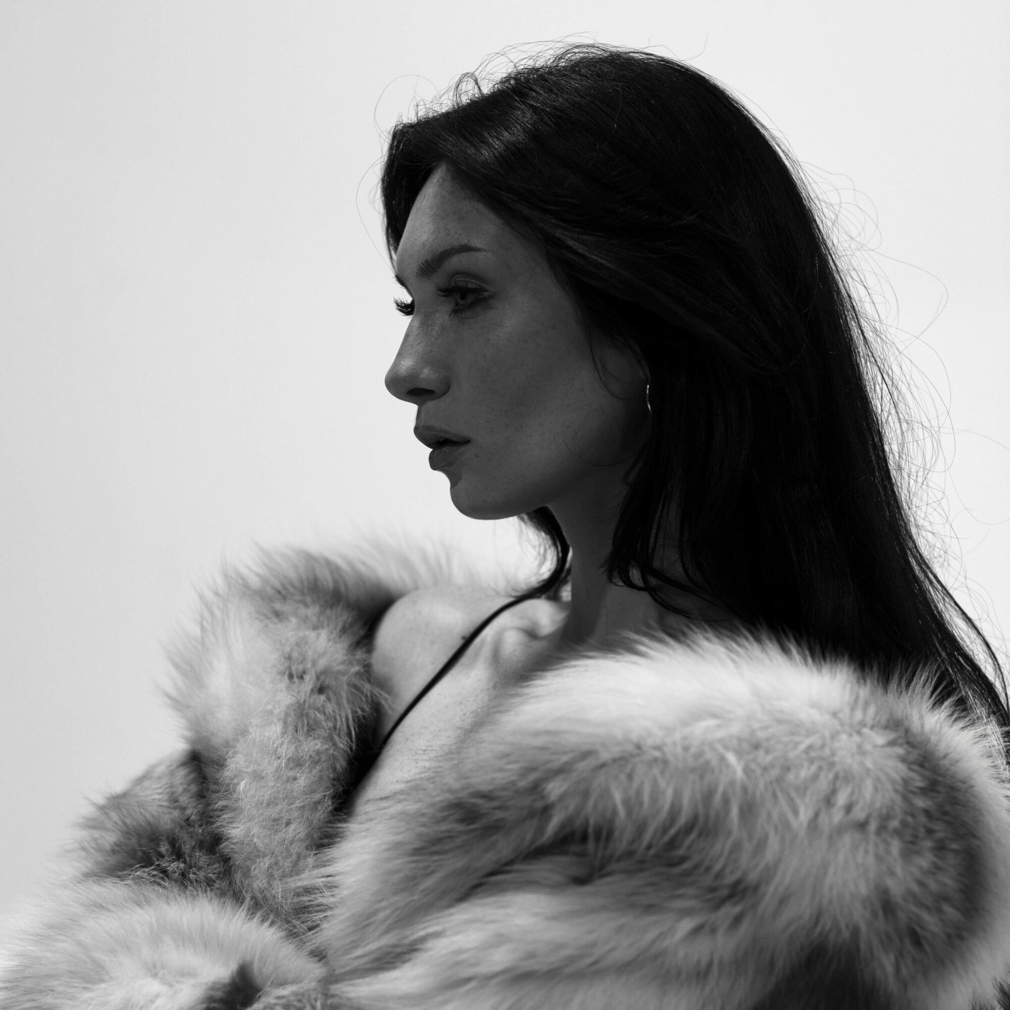Side profile of a woman with long dark hair wearing a fur coat, looking to the left, in black and white.