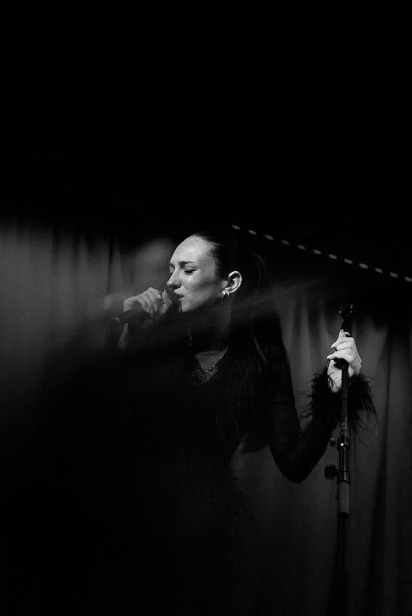 Woman singing into a microphone on stage with a curtain background, holding the microphone stand, in black and white.