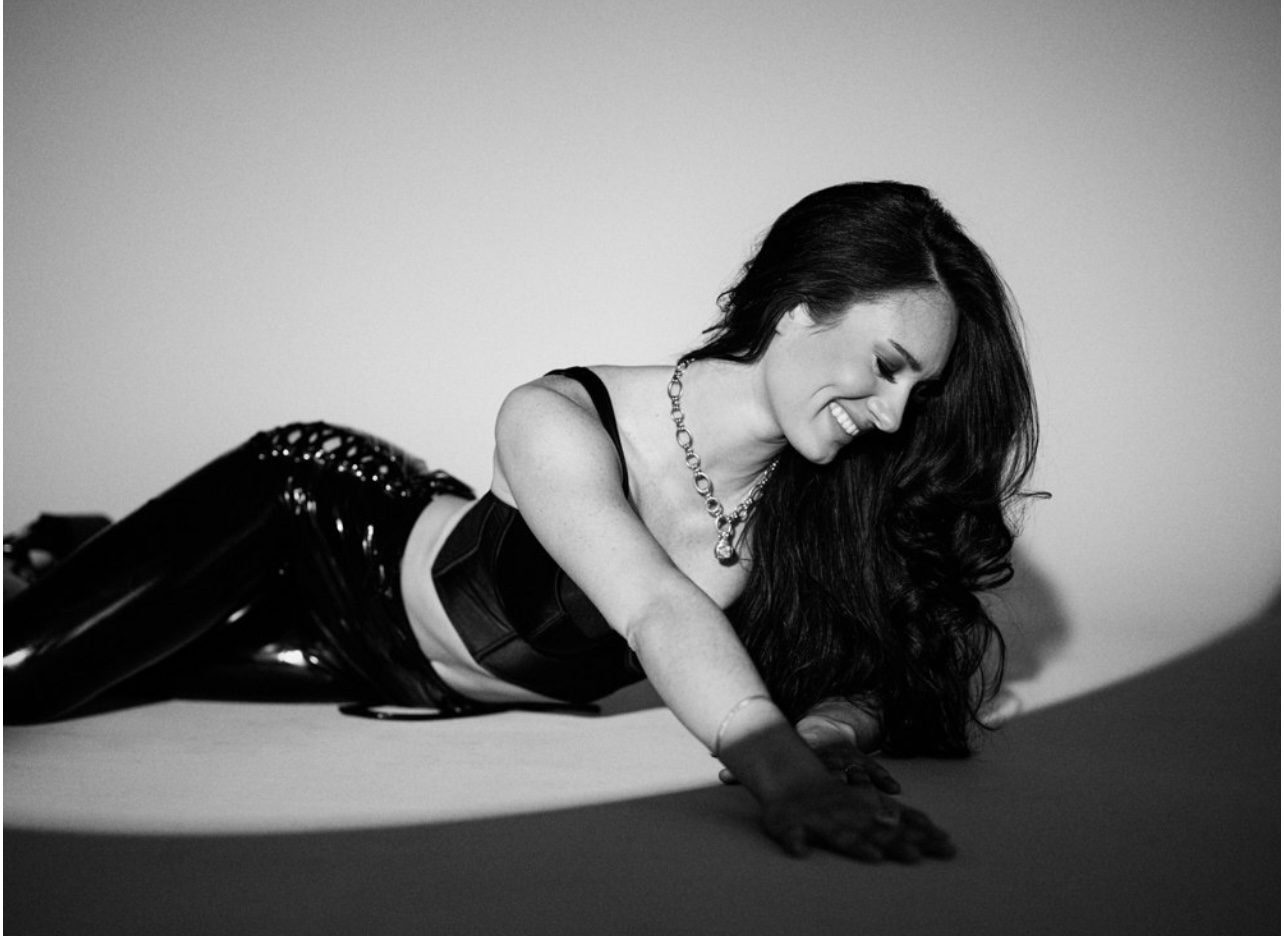 Woman lying on the floor, smiling, wearing a shiny outfit and necklace, with long hair, in black and white.