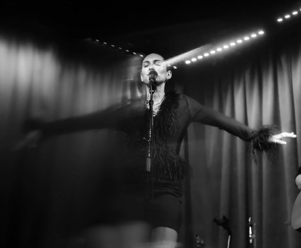 Person singing on stage with arms outstretched, curtains in background, stage lights above, black and white image.