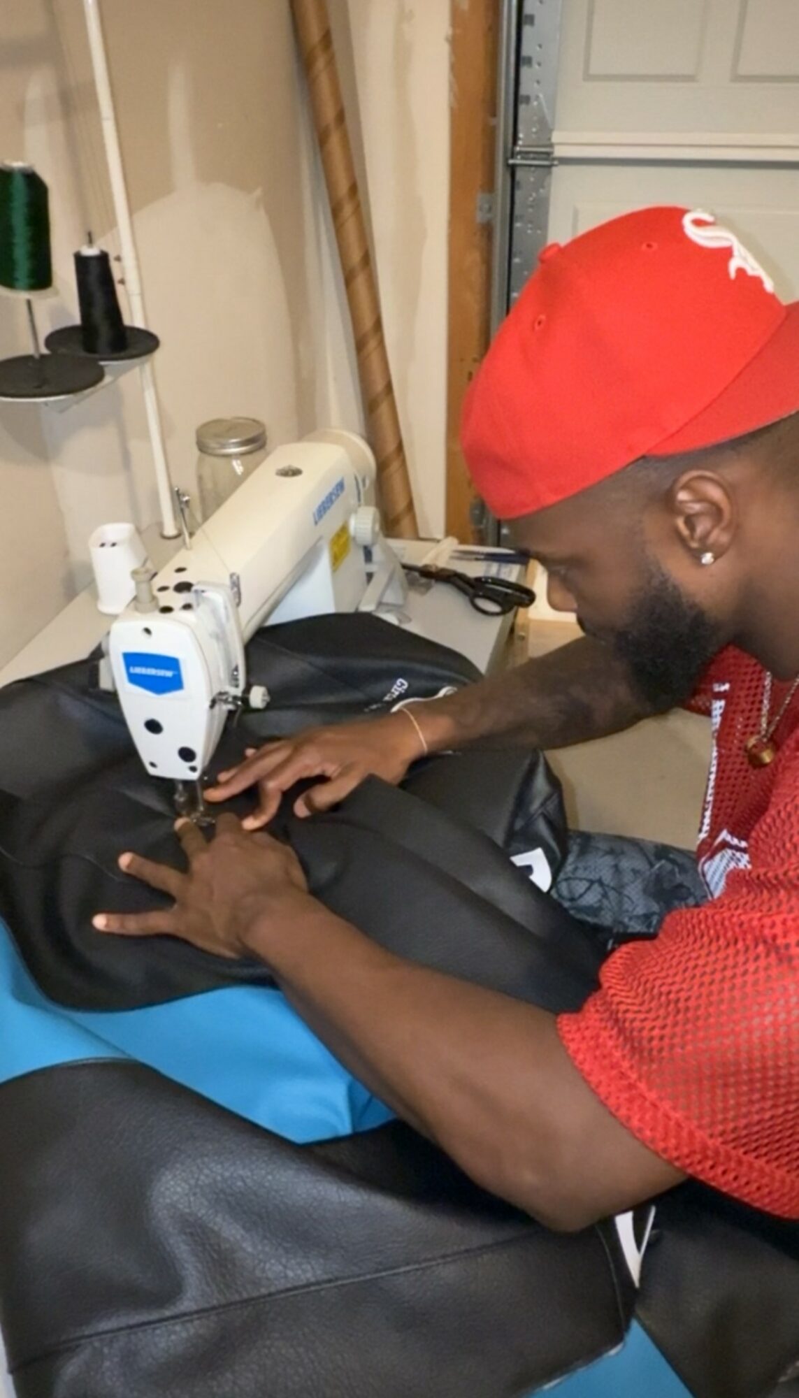 Person sewing fabric with a sewing machine in a workshop, wearing a red head covering and red shirt, focused on work.