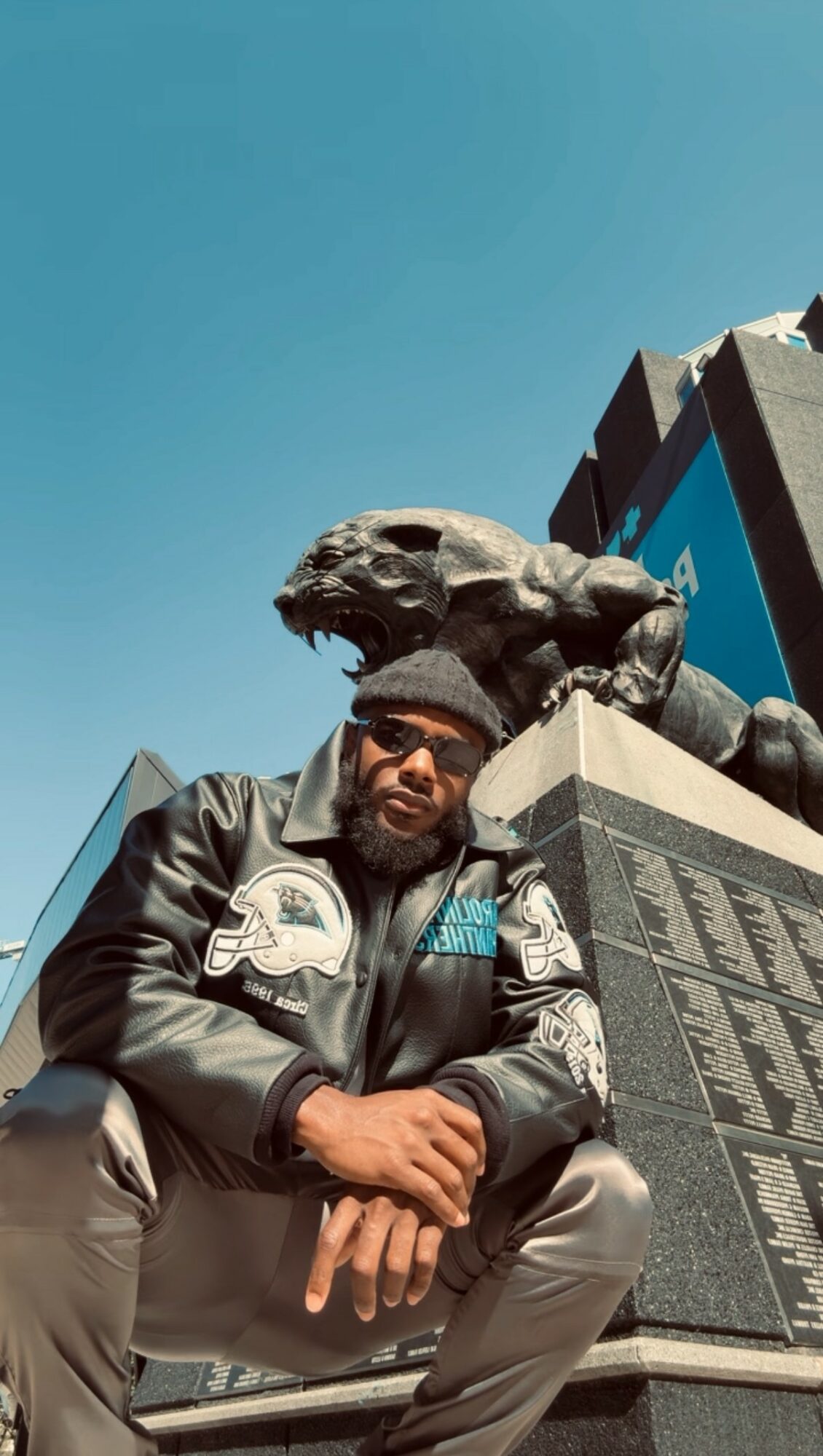 Person wearing sunglasses and a leather jacket with patches, sitting in front of a monument with a lion sculpture on top, under a clear blue sky.
