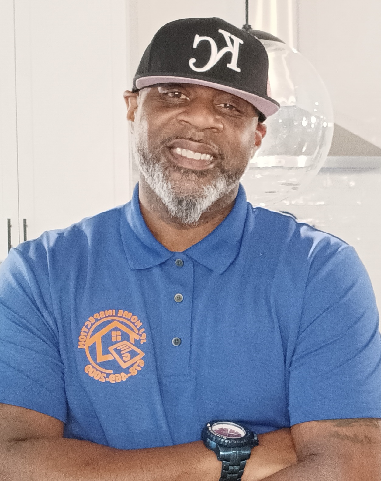 Man with gray beard wearing a black cap and blue polo shirt, smiling, arms crossed, indoors.