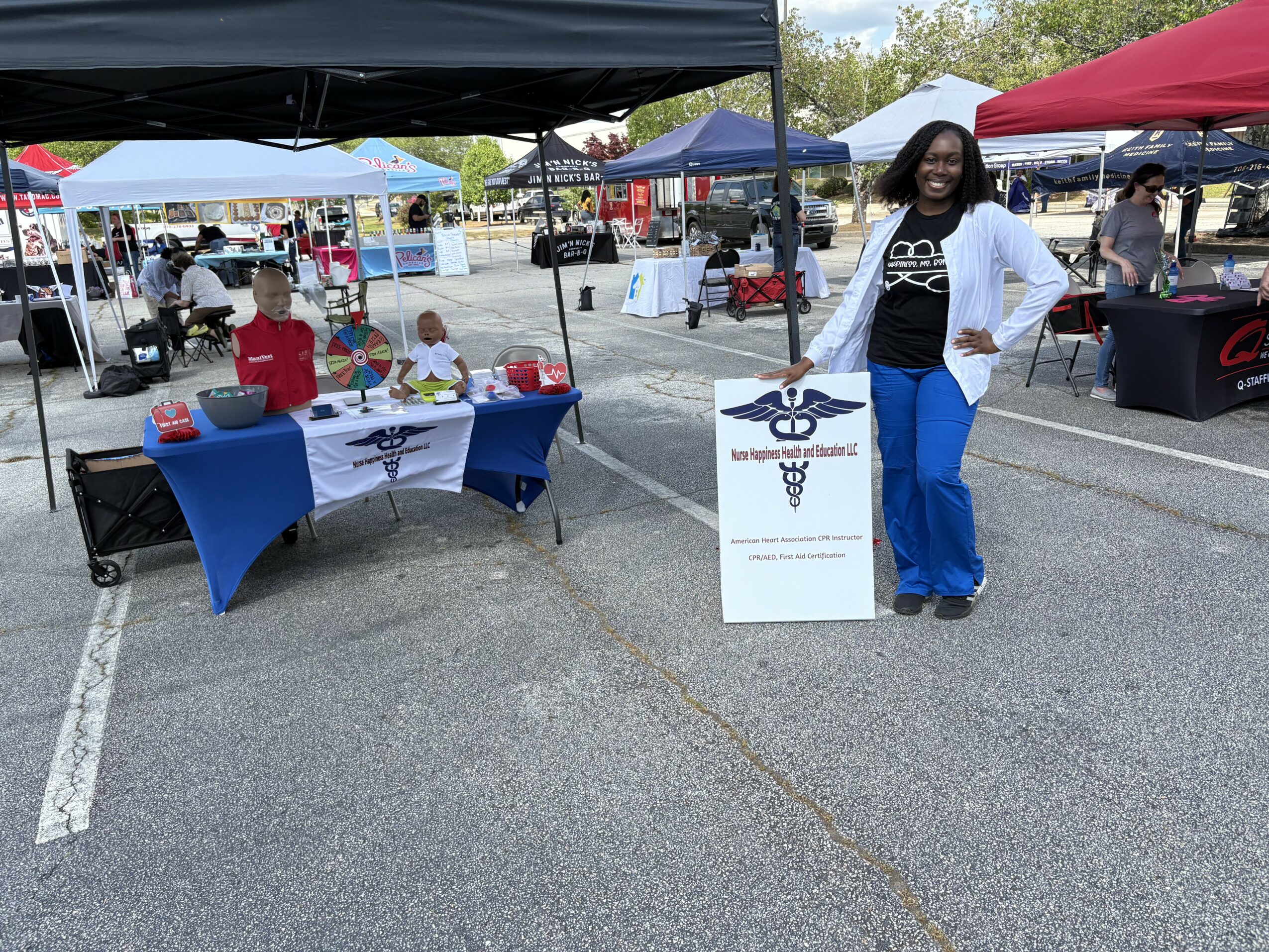 Woman standing next to a sign at an outdoor market, with tents and tables in the background.
