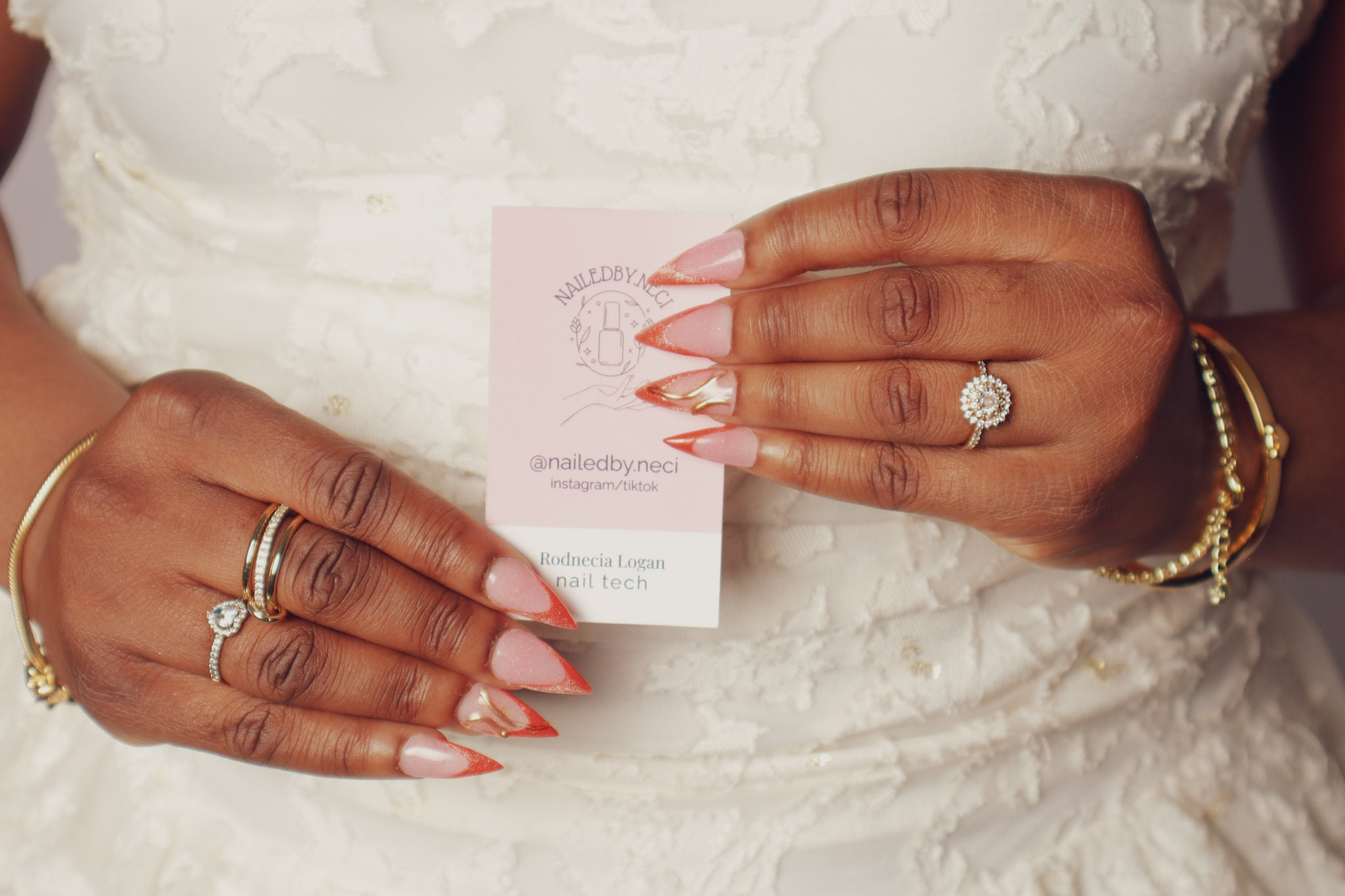 Person holding a business card with both hands, wearing rings and bracelets, in front of a white textured background.