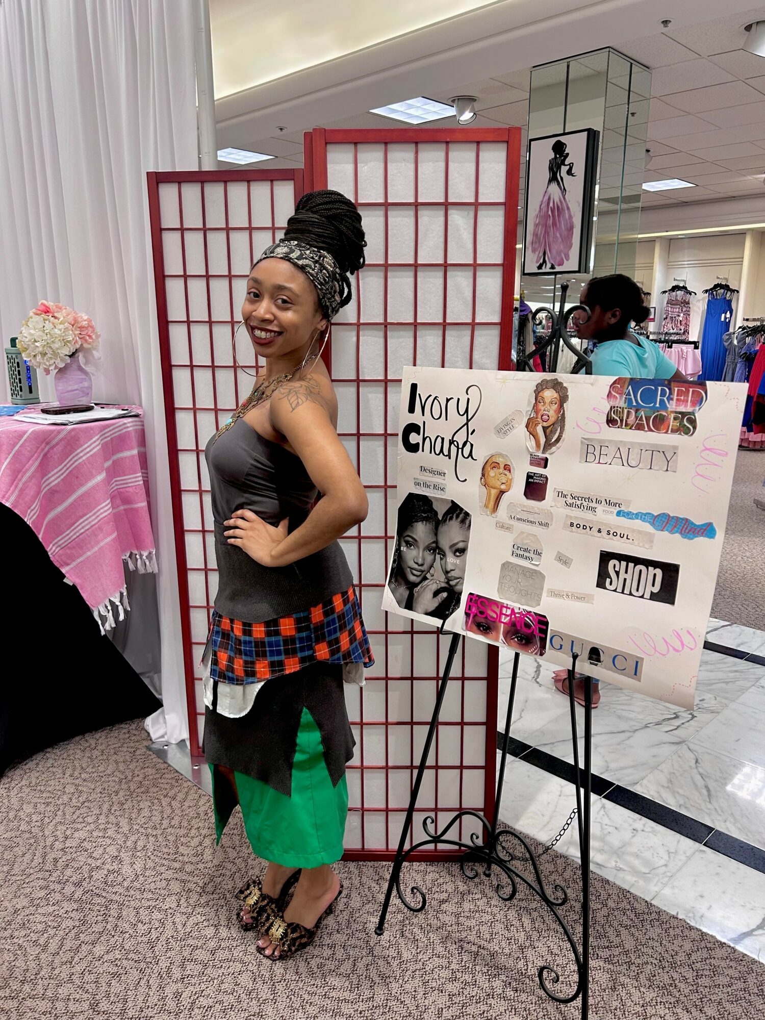 Smiling woman in colorful dress standing next to a display board in a shopping mall, with a pink table and flowers nearby.