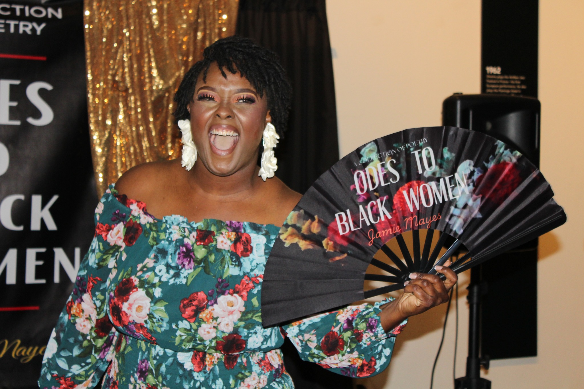 Woman smiling and holding a fan with text, wearing a floral off-shoulder dress, in front of a black and gold backdrop.