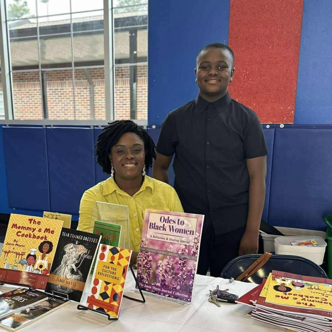 Two people standing behind a table with books, one wearing a yellow shirt and the other in a black shirt, in a colorful indoor setting.