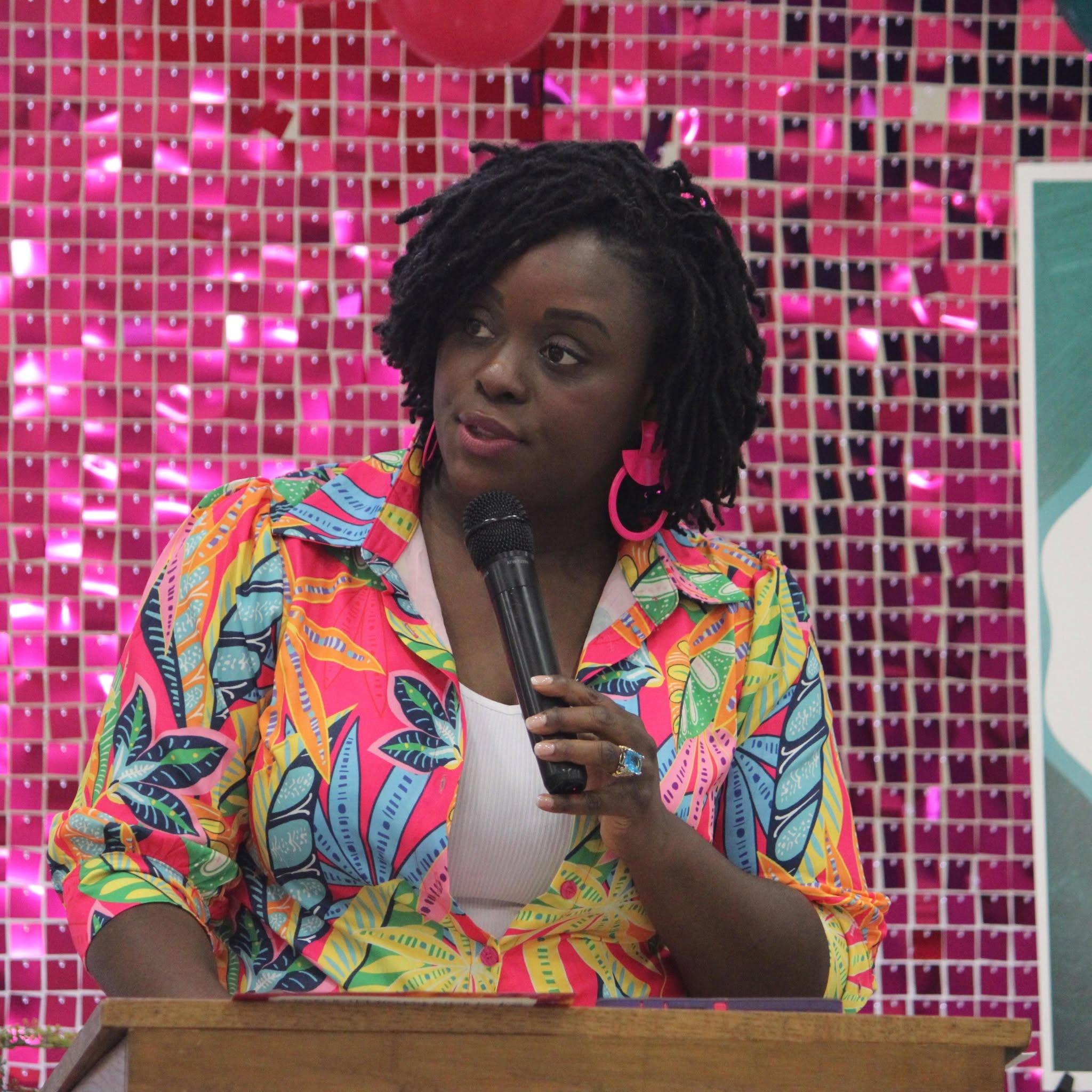 Woman with short curly hair holding a microphone, wearing a colorful patterned shirt, standing in front of a pink mosaic background.