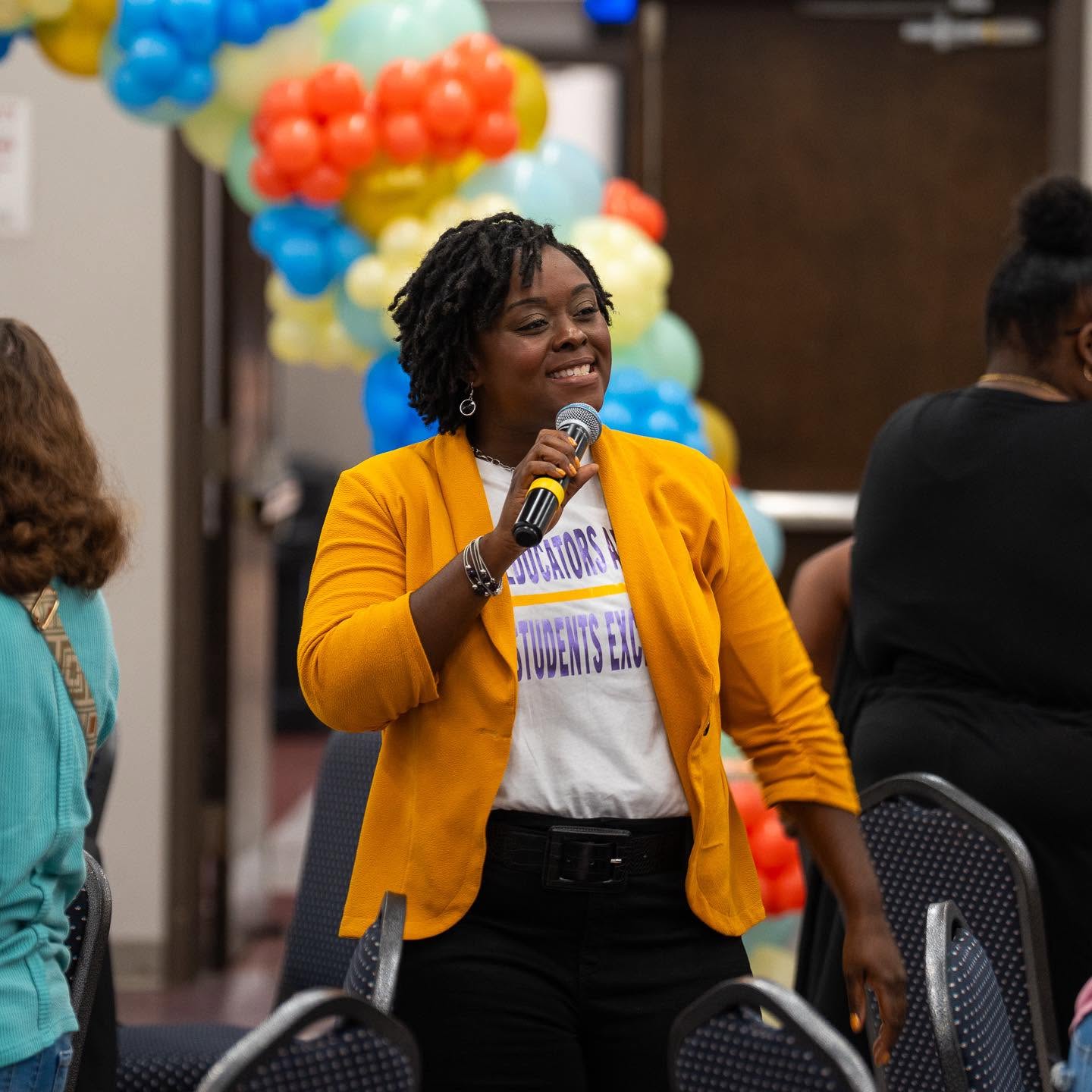 Woman speaking into microphone at event, smiling, wearing yellow blazer and white shirt, with colorful balloon decorations behind her.