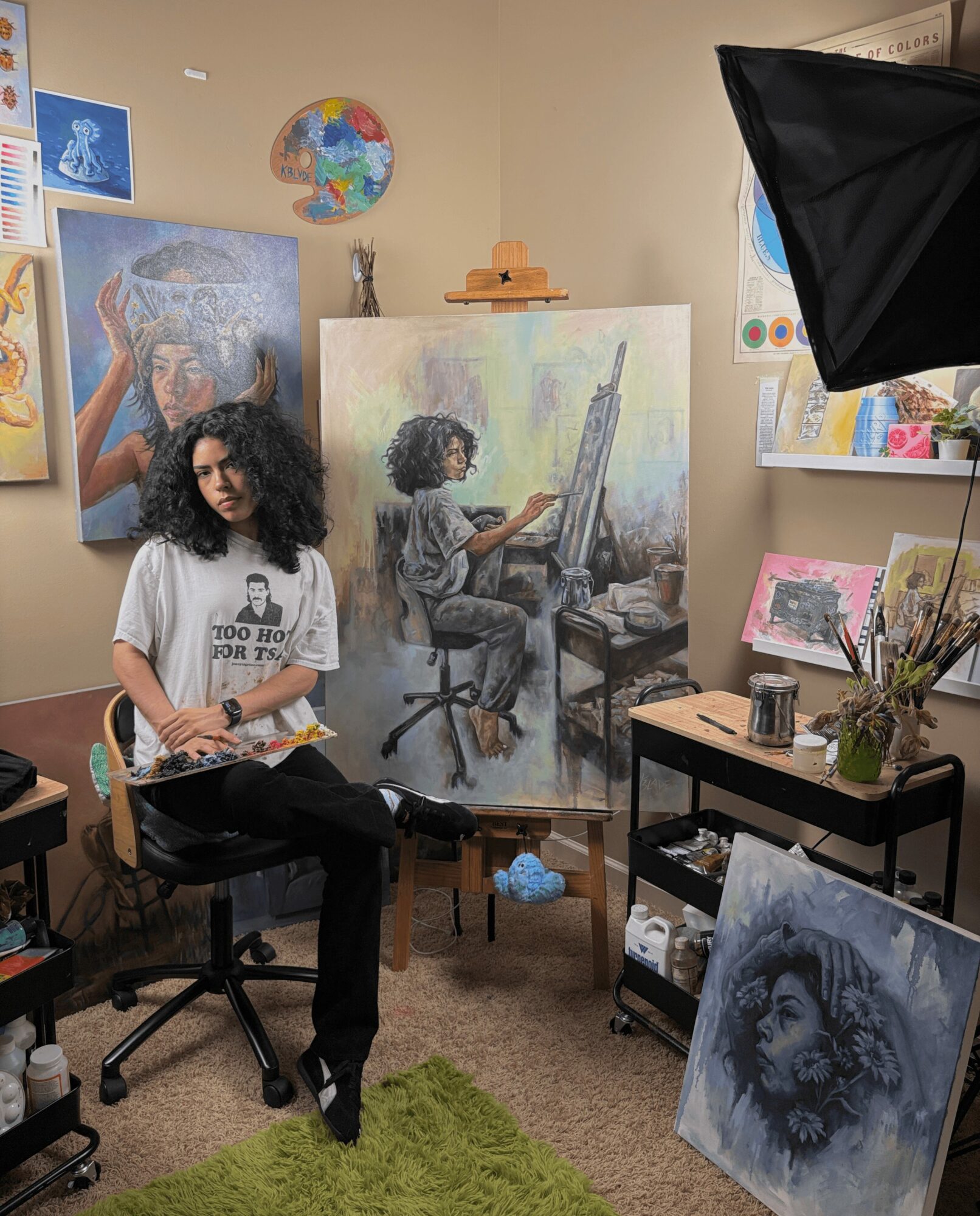 Person with curly hair holding a book in an art studio with paintings and art supplies, green rug on floor.