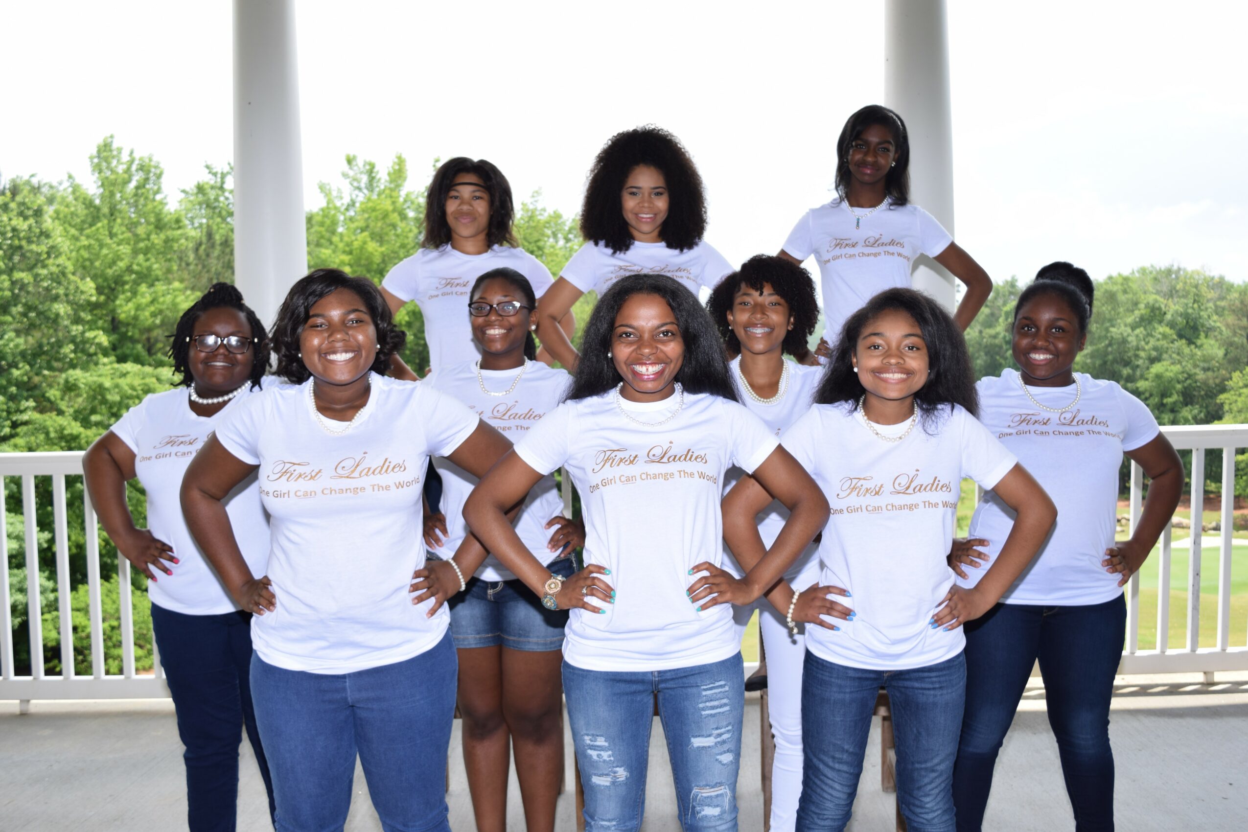 Group of nine young girls standing outdoors on a porch, smiling with hands on hips, wearing white T-shirts and jeans.