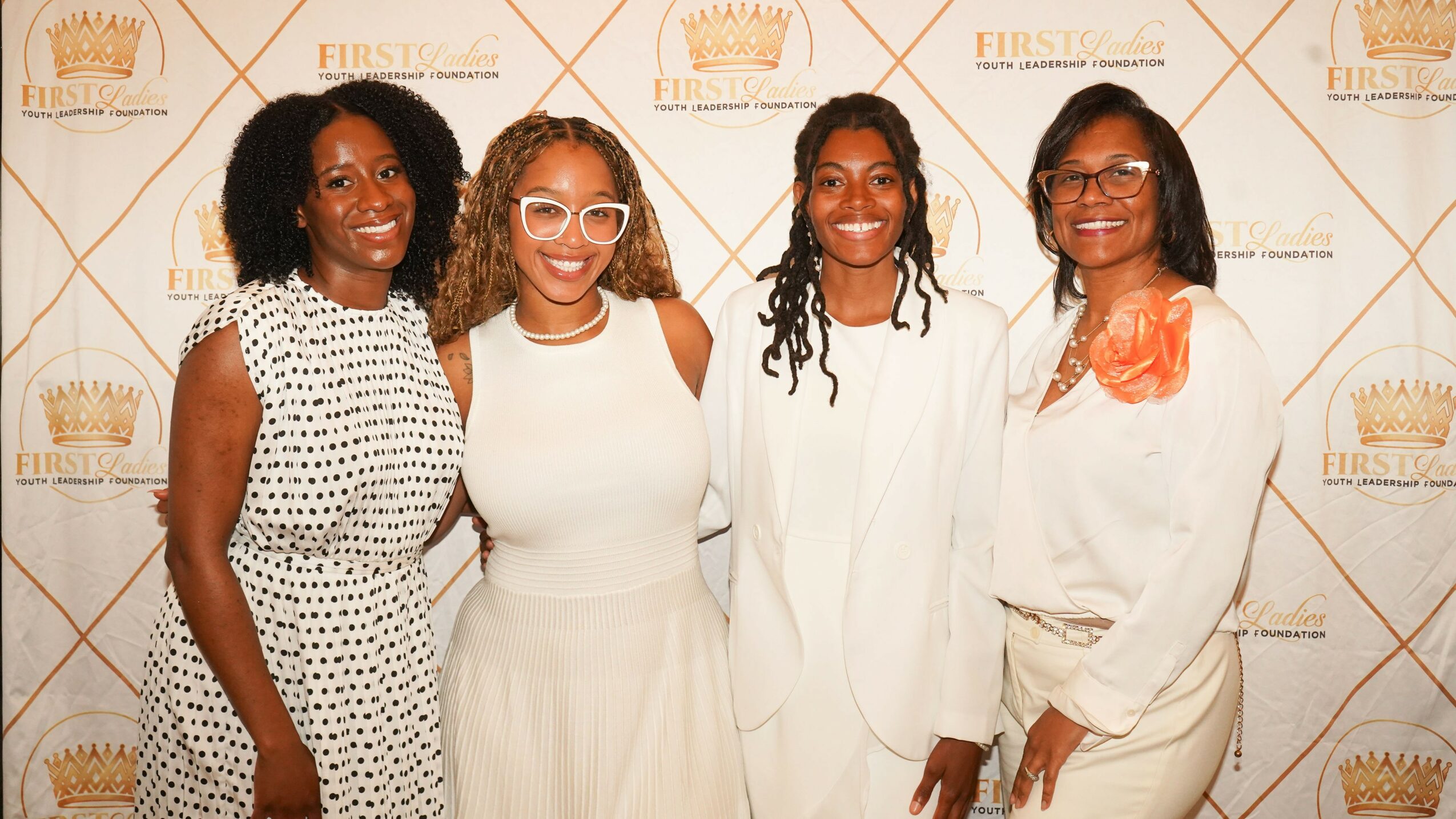 Four women standing together in front of a patterned backdrop, smiling and dressed in white and patterned clothing.