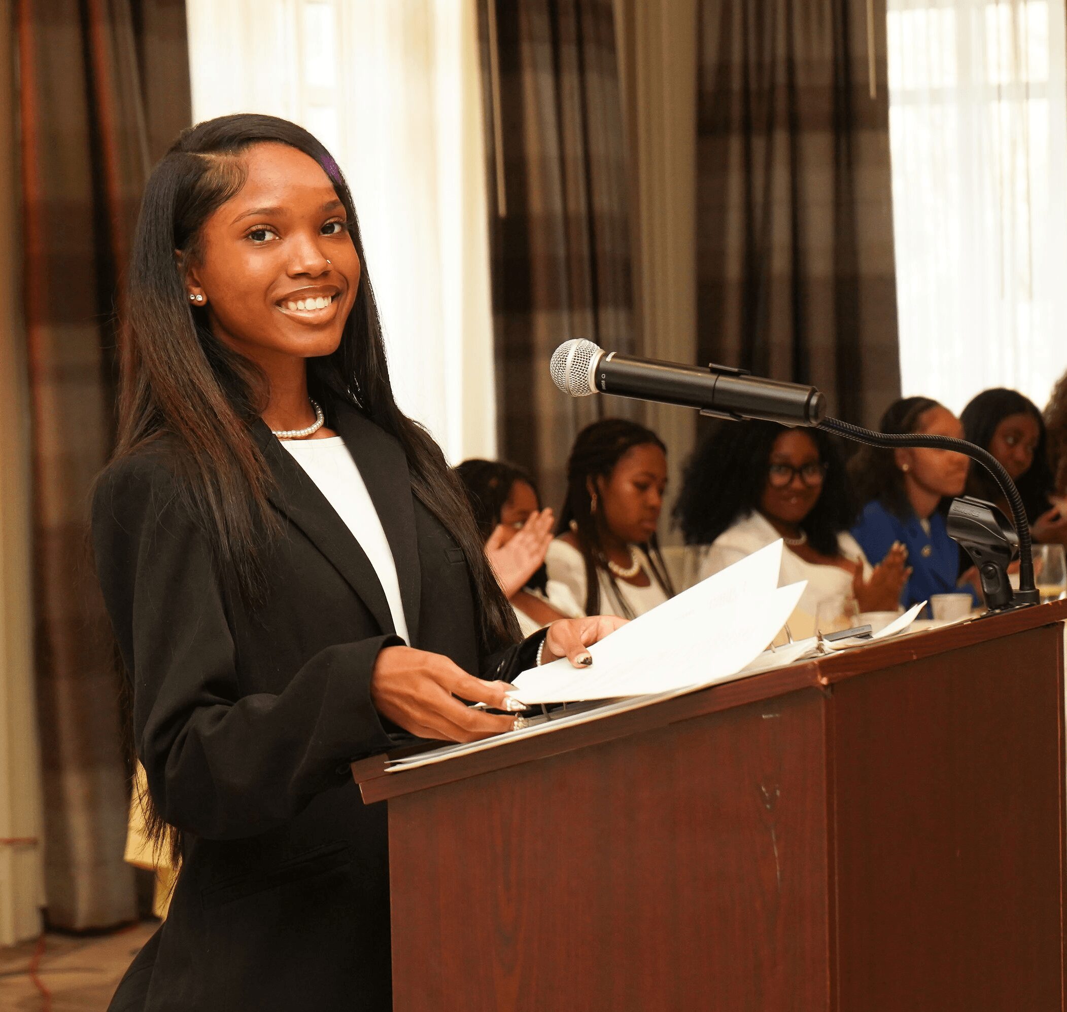 Young woman speaking at a podium during a formal event, with a group of people seated behind her at a long table.