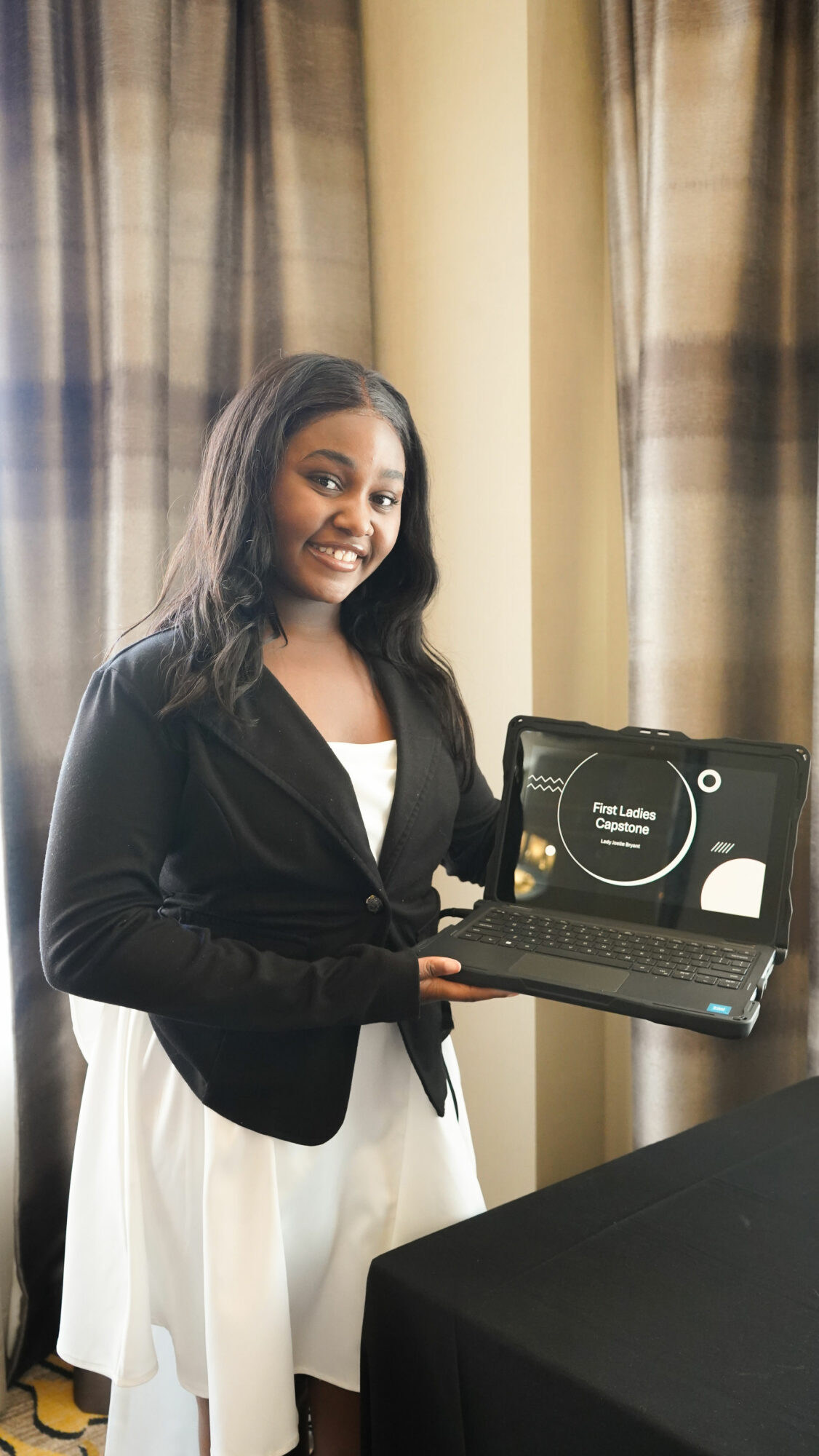 Woman holding a laptop with a presentation slide, standing indoors near curtains, smiling, wearing a black blazer and white dress.