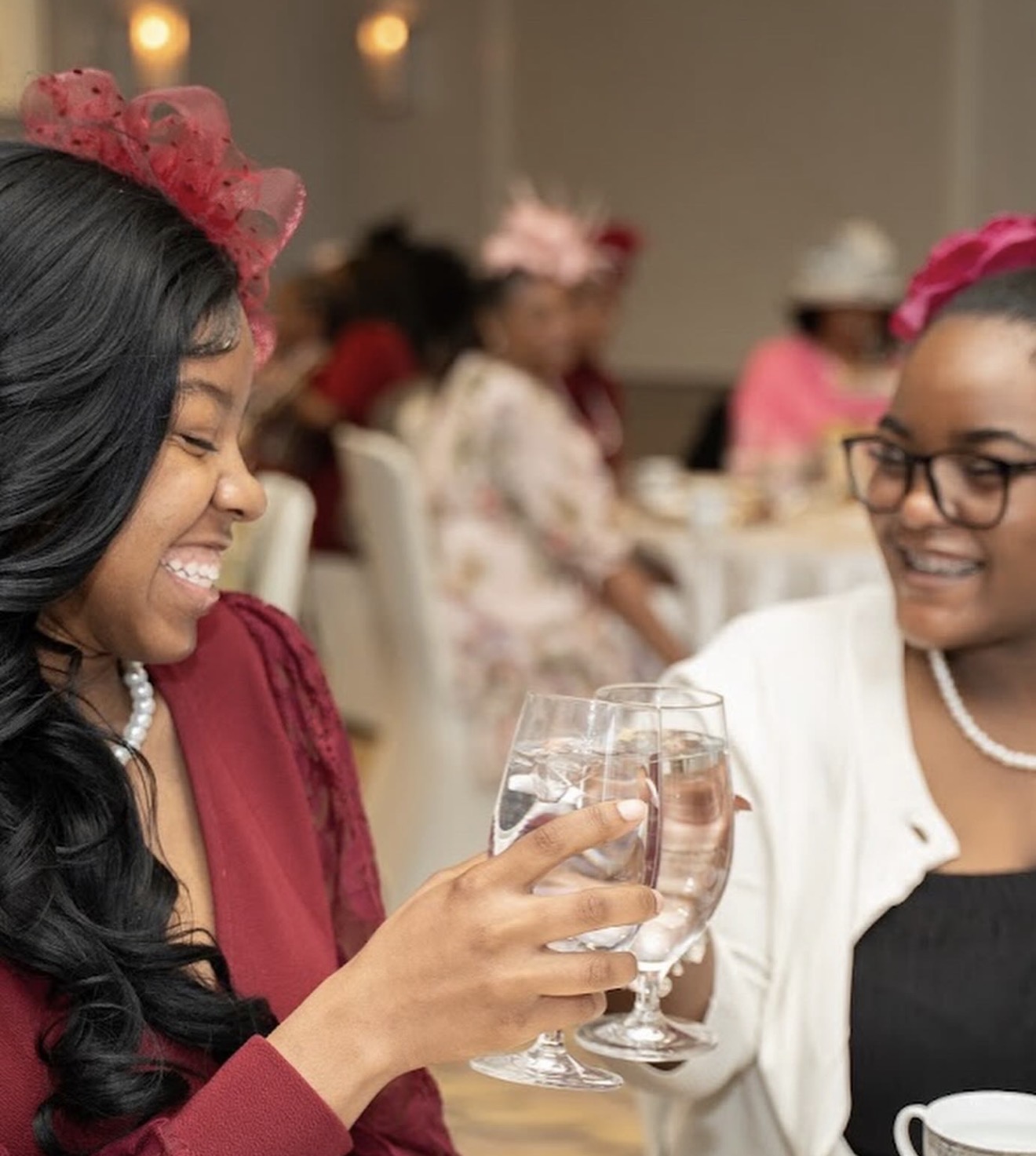 Two women smiling and clinking glasses at a celebration, with other guests in the background wearing hats and festive attire.