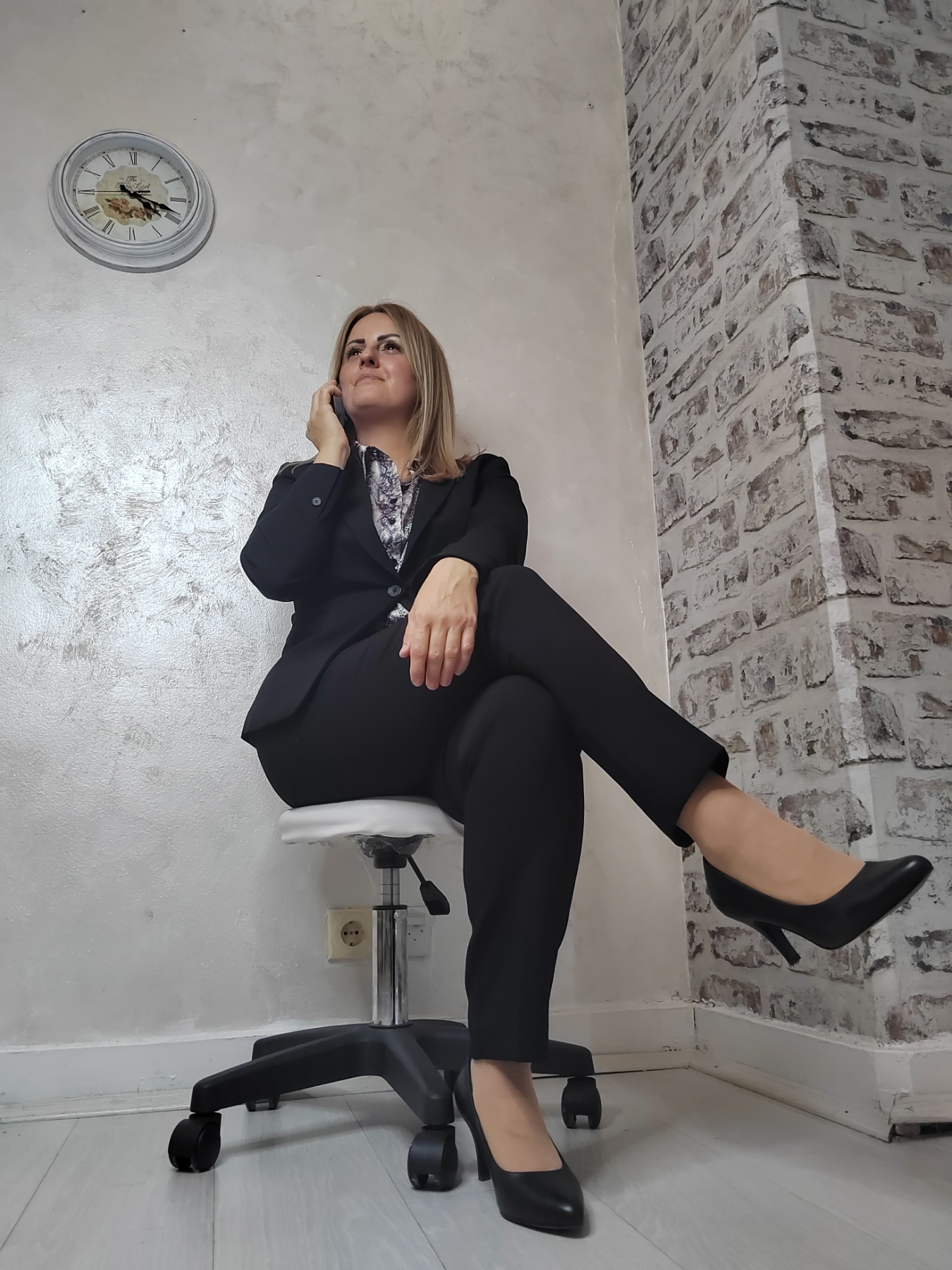 Woman in formal attire sitting on a chair with one leg crossed, looking upward, against a wall with a clock and brick pattern.