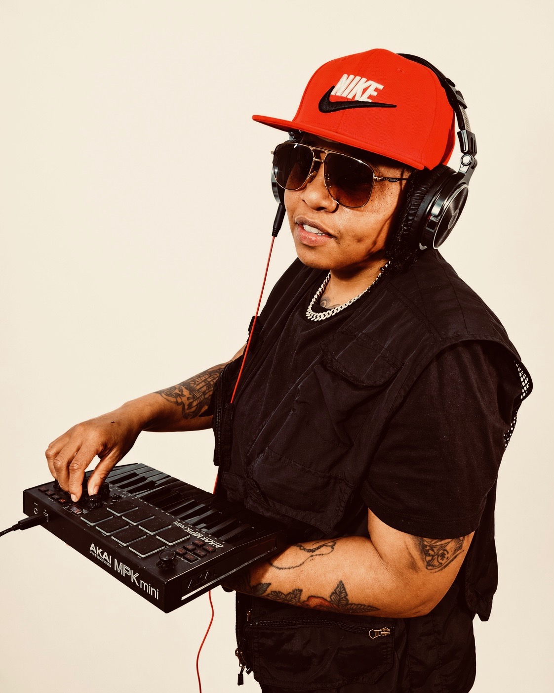Person wearing sunglasses, red Nike cap, headphones, black vest, holding music equipment, smiling, against a plain background.