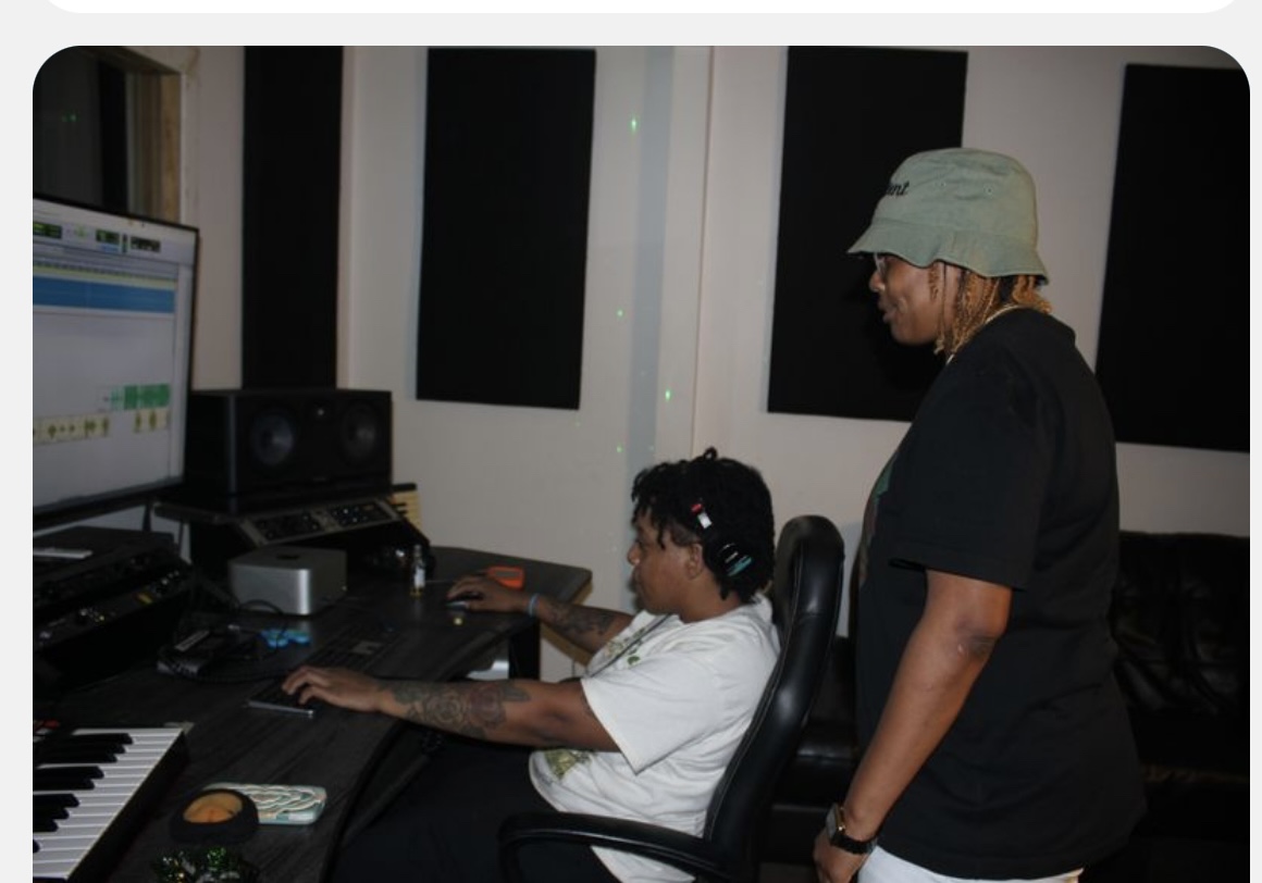 Two people in a recording studio, one sitting at a computer and the other standing nearby, with soundproof panels on the wall.
