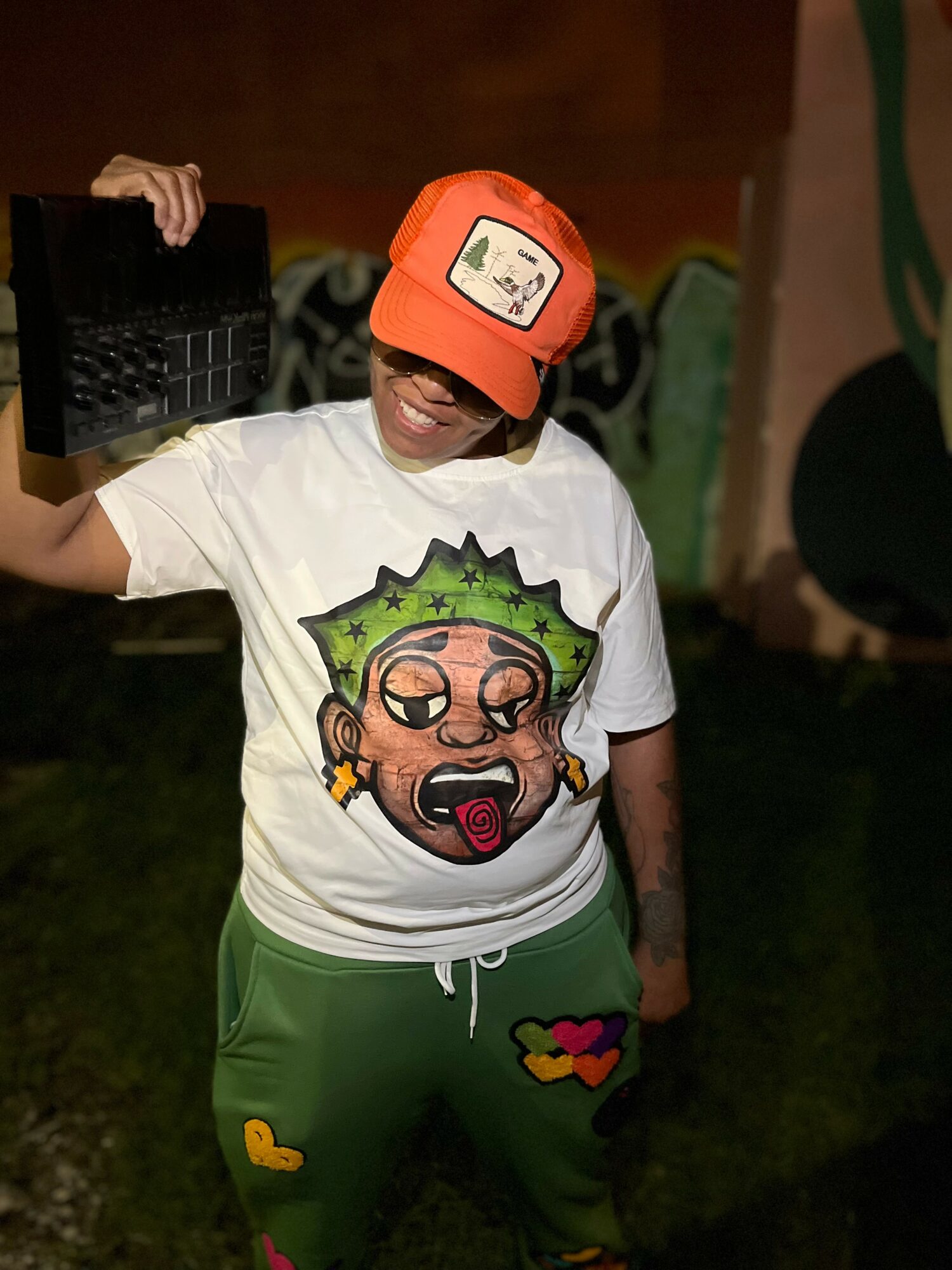 Child wearing a white T-shirt with a cartoon face, green pants with colorful patches, and an orange cap, holding a black object.