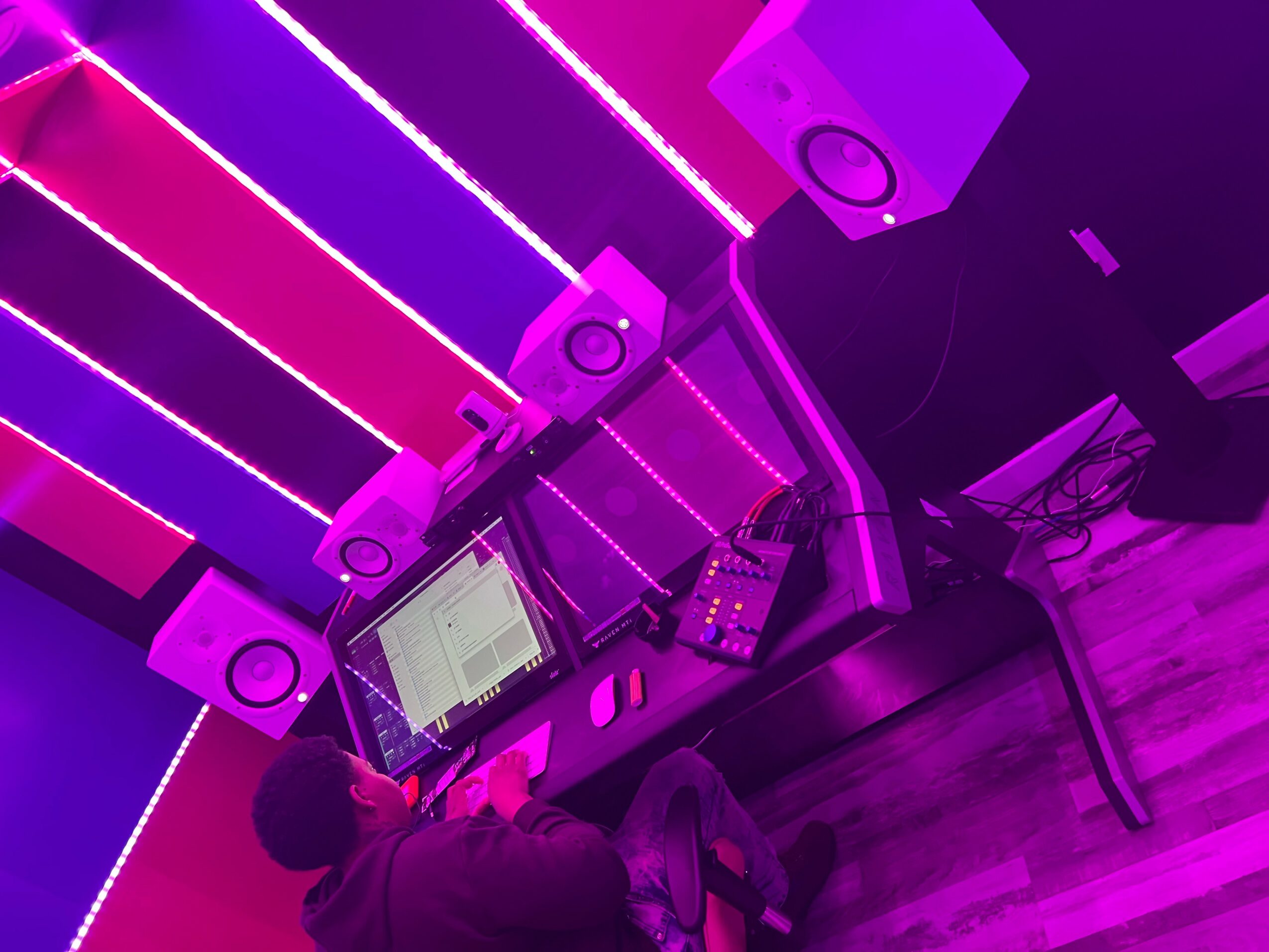 DJ setup with speakers, laptop, and mixer illuminated by pink and purple neon lights, on a wooden floor.
