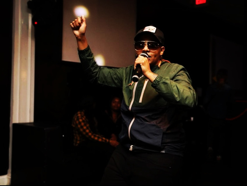 Person wearing sunglasses and a cap, holding a microphone, raising a fist, in a dark indoor setting.