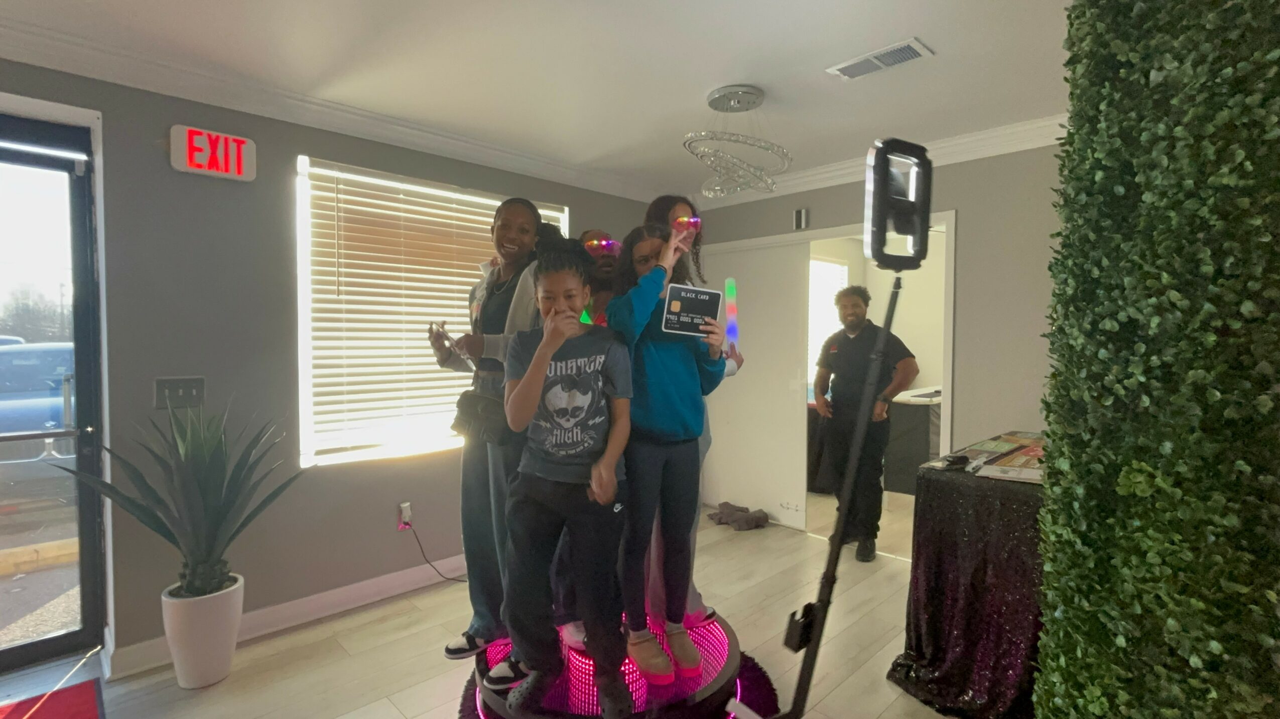 Group of children standing on a pink trampoline indoors, with a woman and a man nearby, in a room with windows and a potted plant.