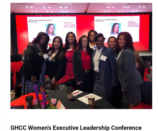 Group of women standing together at a conference, smiling, with screens displaying a woman’s face in the background.