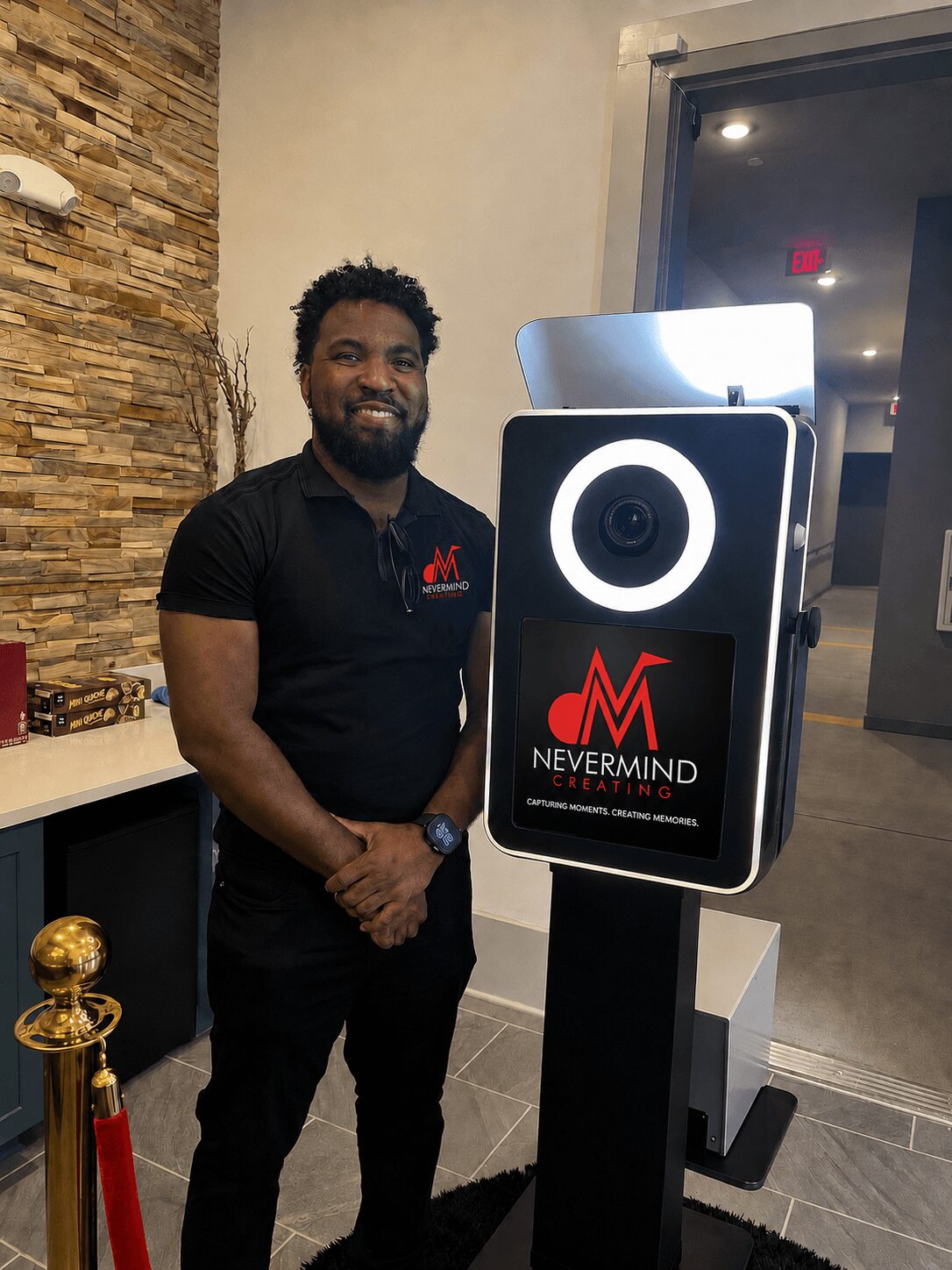 Man with beard and black shirt standing indoors next to a photo booth with a Nevermind logo, smiling at the camera.