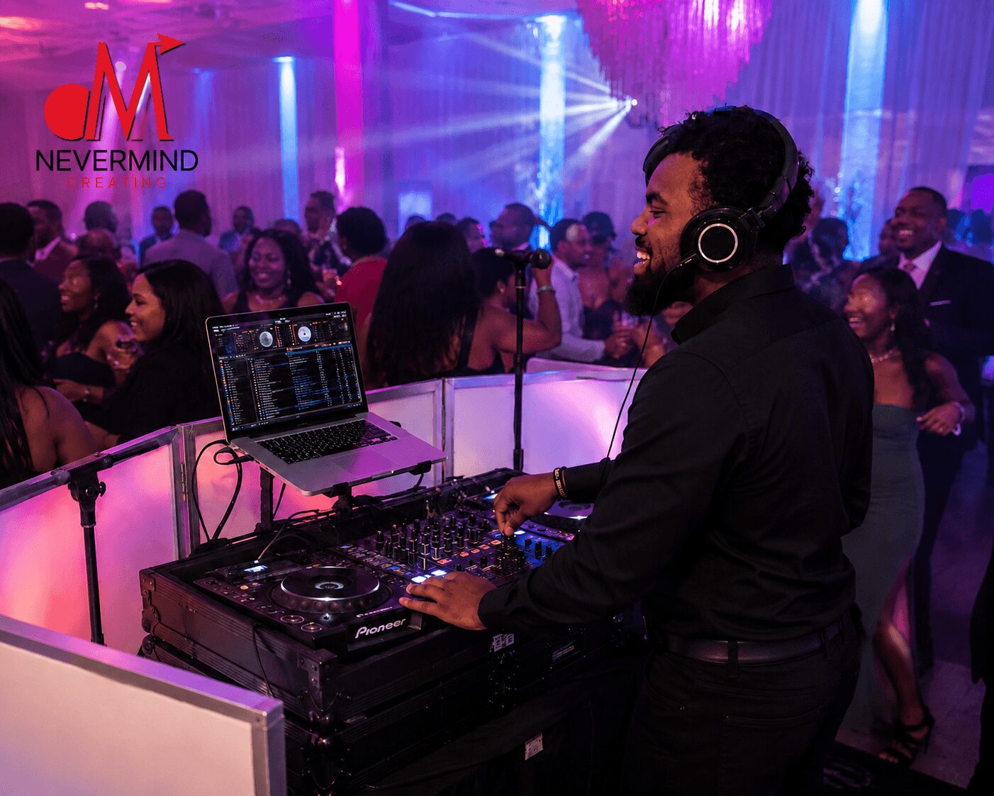 DJ with headphones operating equipment at a lively party with colorful lighting and a crowd of people dancing.