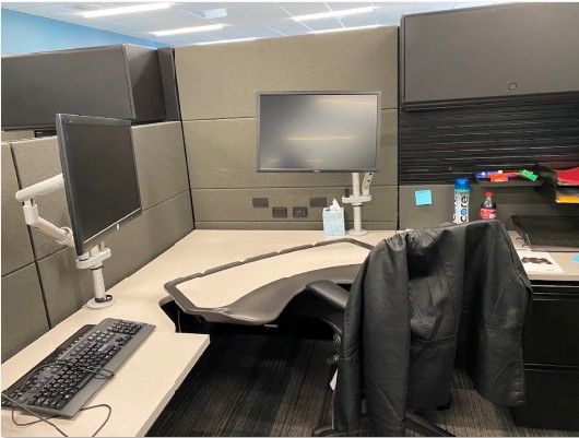 Office workspace with dual monitors, keyboard, and a chair with a jacket draped over it, in a cubicle setting.