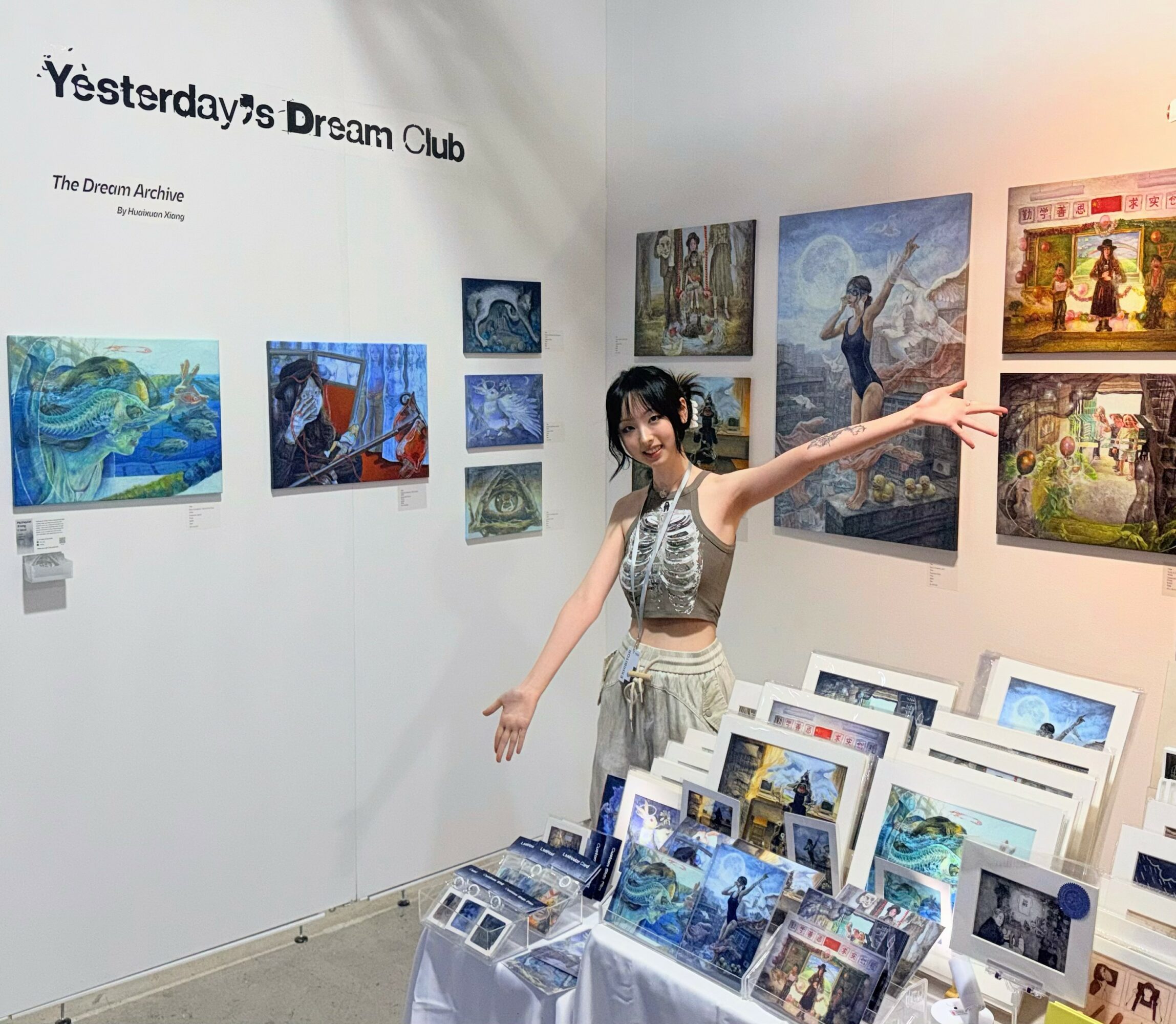 Young woman in a patterned top and skirt stands in an art gallery, gesturing towards paintings on the wall.