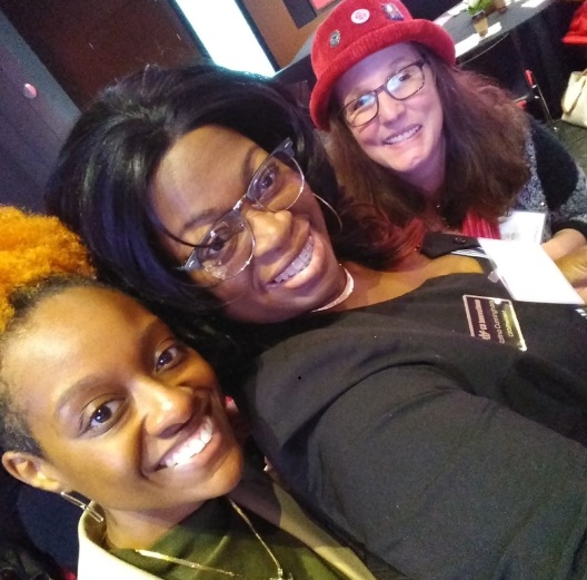 Three women smiling and posing for a selfie indoors, one wearing glasses and a red hat, others with glasses and curly hair.