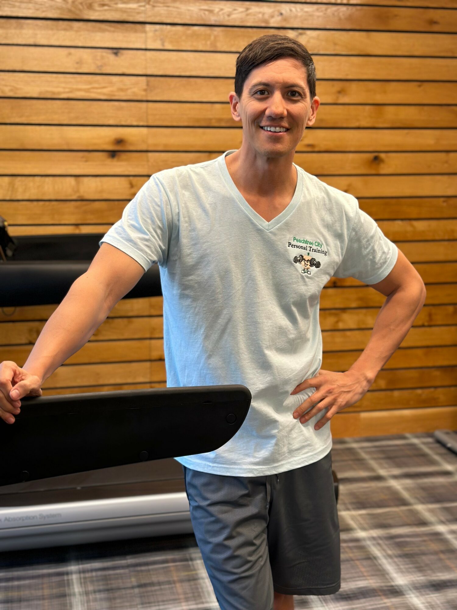 Young man smiling, standing indoors with wooden wall background, wearing a light gray T-shirt and dark shorts, leaning on a treadmill.