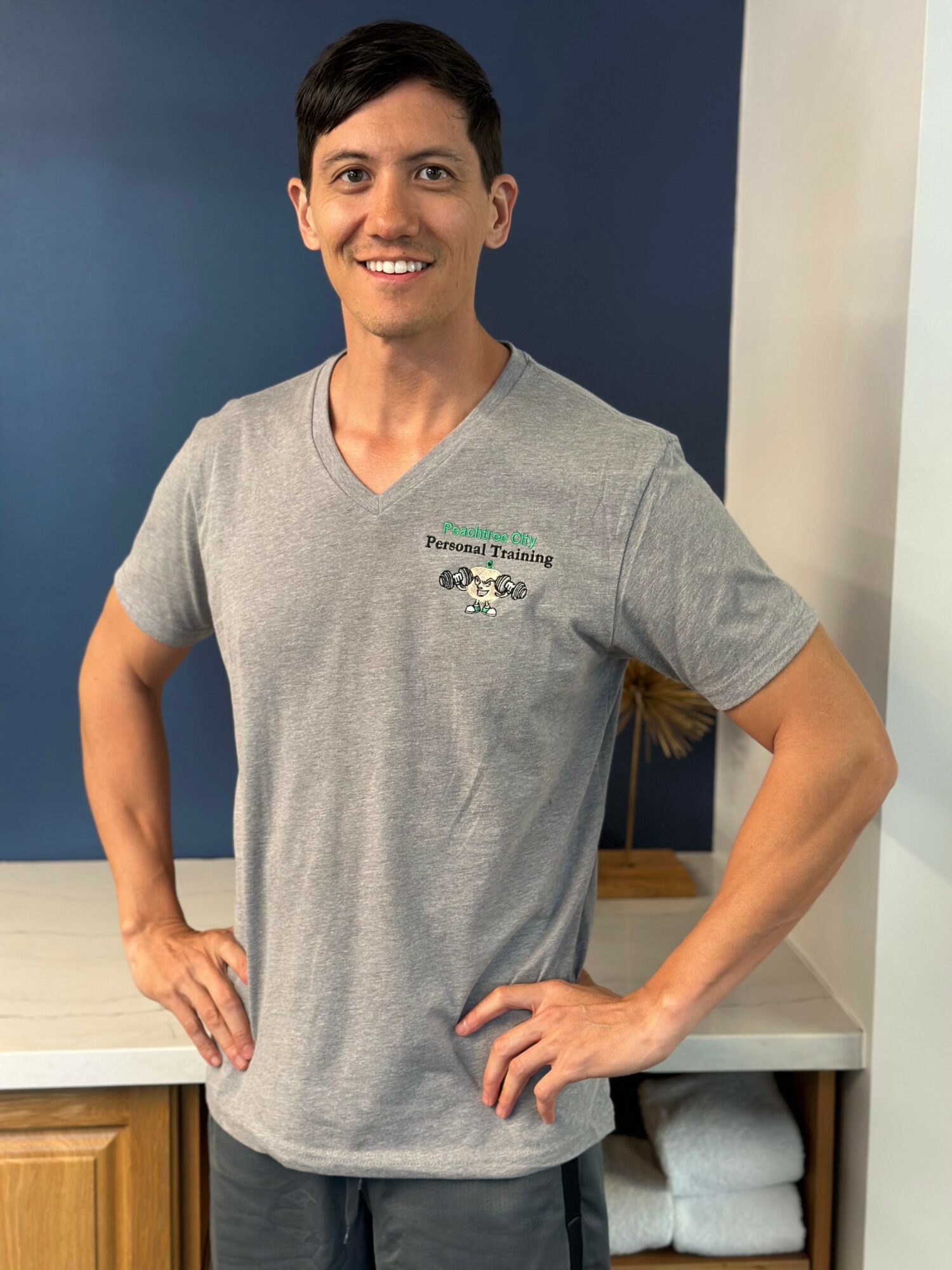 Person with short dark hair smiling, wearing a gray V-neck T-shirt, standing indoors with hands on hips.