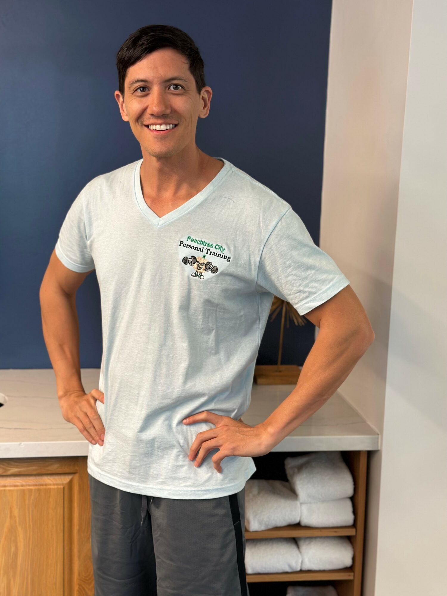 Smiling person with short dark hair wearing a white T-shirt standing indoors with hands on hips, next to a shelf with folded towels.