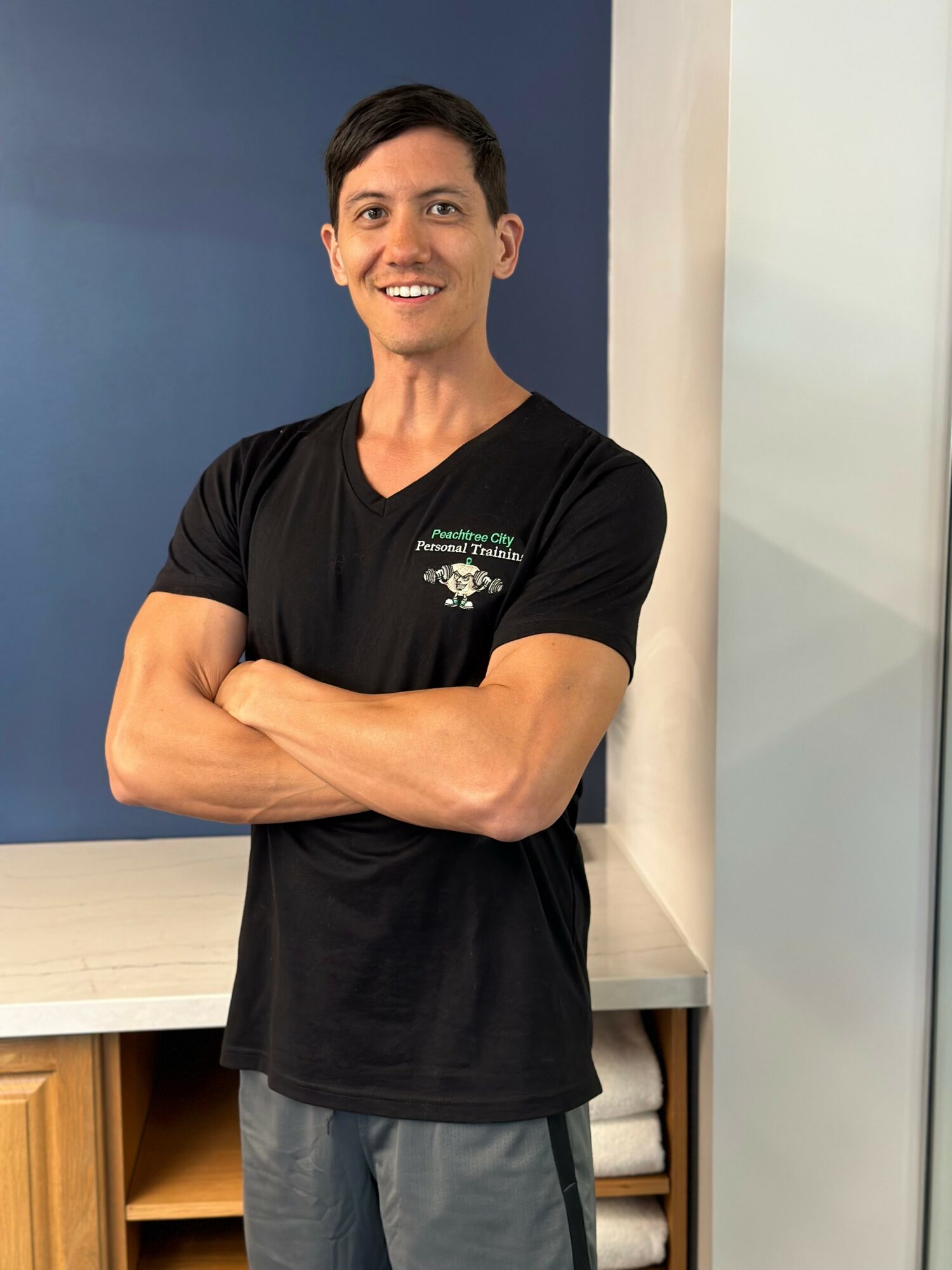 Smiling man with crossed arms standing indoors near a white wall and blue background, wearing a black T-shirt.