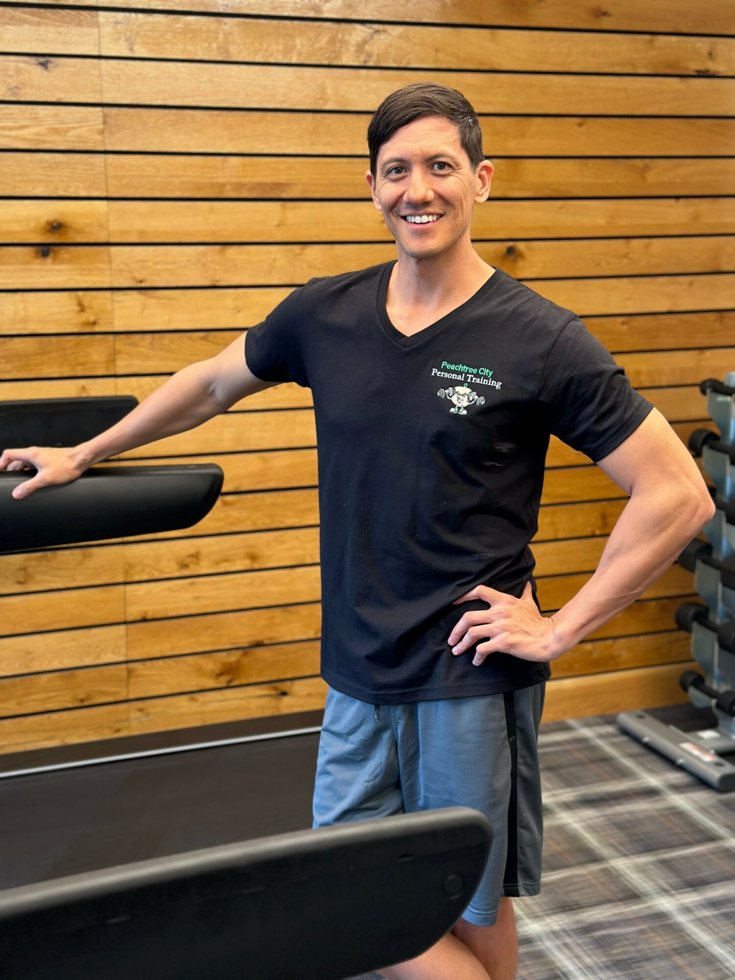 Smiling person standing in a gym, wearing a black T-shirt and blue shorts, with a wooden wall background.